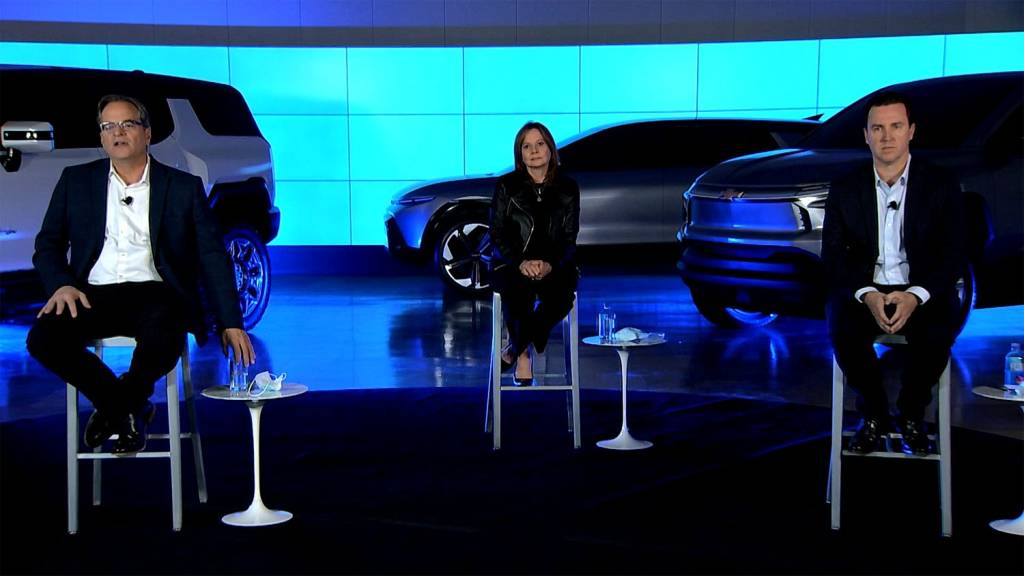 Reunião da GM com três pessoas. No meio, a CEO da marca, Mary Barra. Atrás deles alguns modelos da marca
