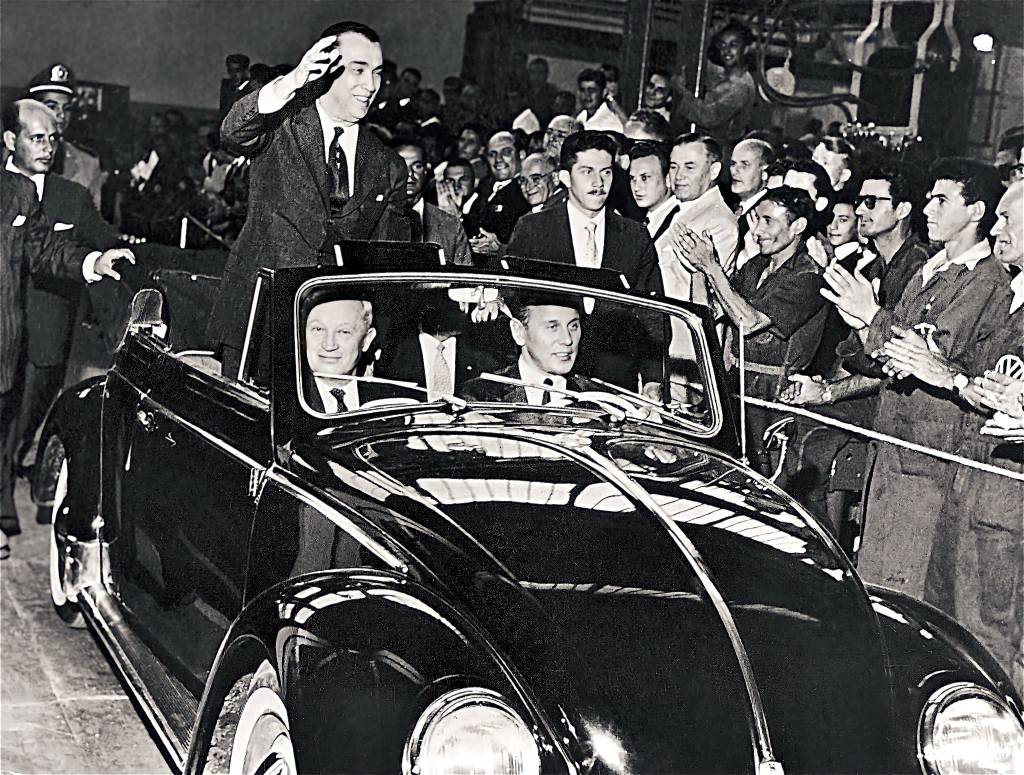 Juscelino Kubitschek acenando num Fusca conversível, conduzido por Henrich Nordhoff, presidente do grupo Volkswagen, no banco de passageiros, e Wilhelm Shultz-Wenk, presidente da Volkswagen no Brasil, ao volante