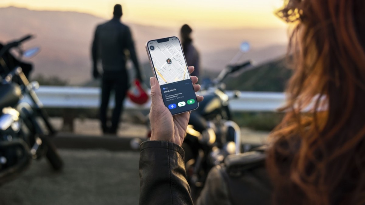 Mão de uma pessoa com jaqueta de couro segurando um smartphone com mapa e informações de contato, com motociclistas e motos desfocados ao fundo em um pôr do sol