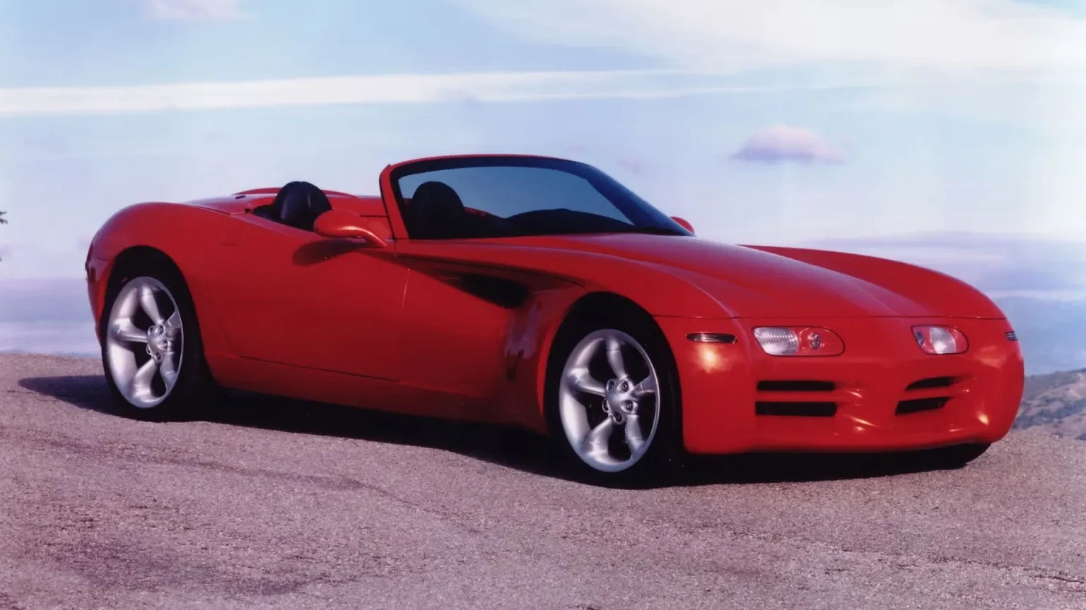 Carro esportivo conversível vermelho brilhante, com capota abaixada, estacionado em uma estrada asfaltada. O veículo tem rodas prateadas e design aerodinâmico. Ao fundo, um céu azul claro com nuvens esparsas e uma paisagem montanhosa desfocada