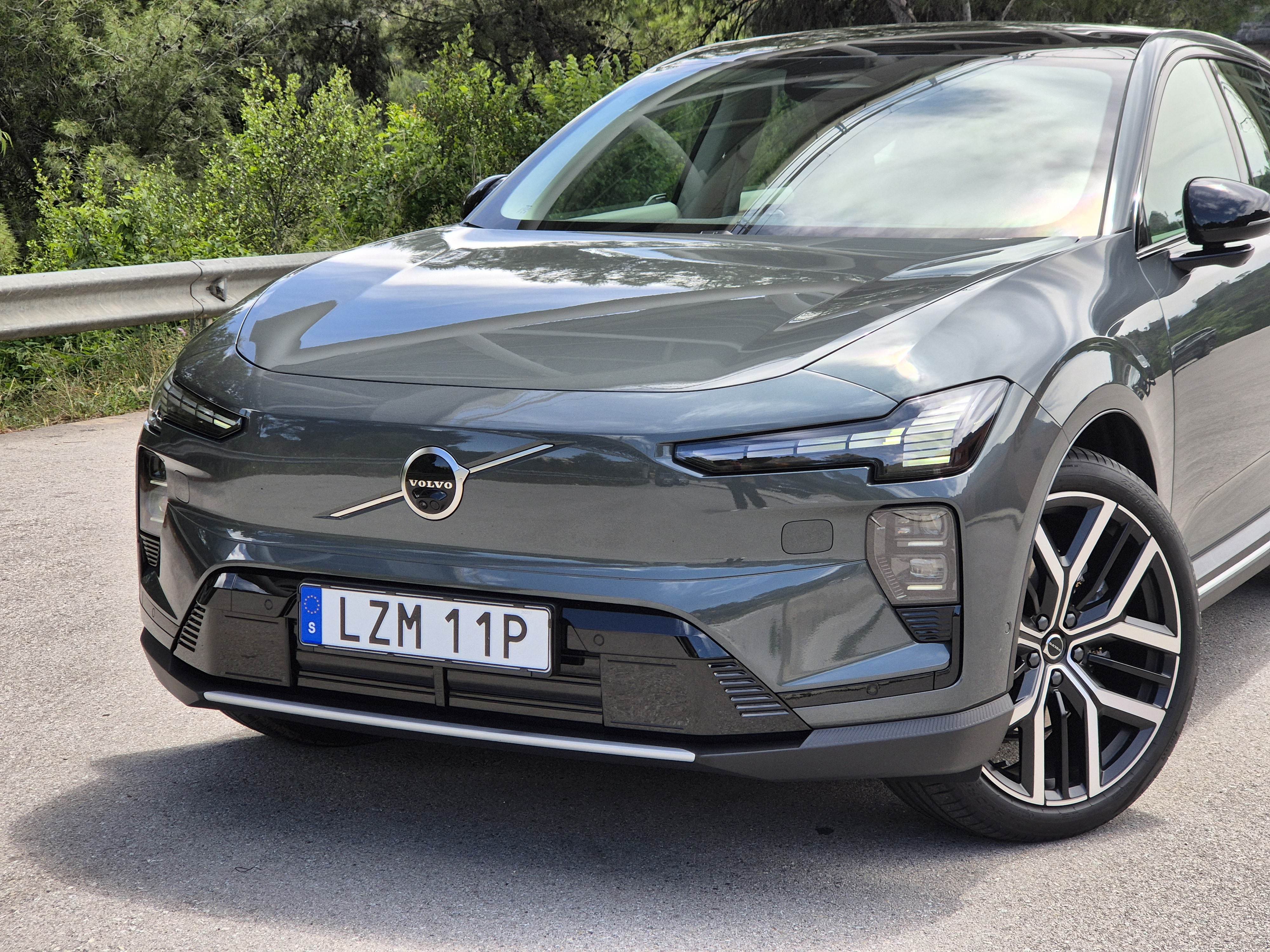 Frente de um Volvo EX60 cinza escuro, com faróis e luzes diurnas acesas, estacionado em uma estrada asfaltada com vegetação ao fundo