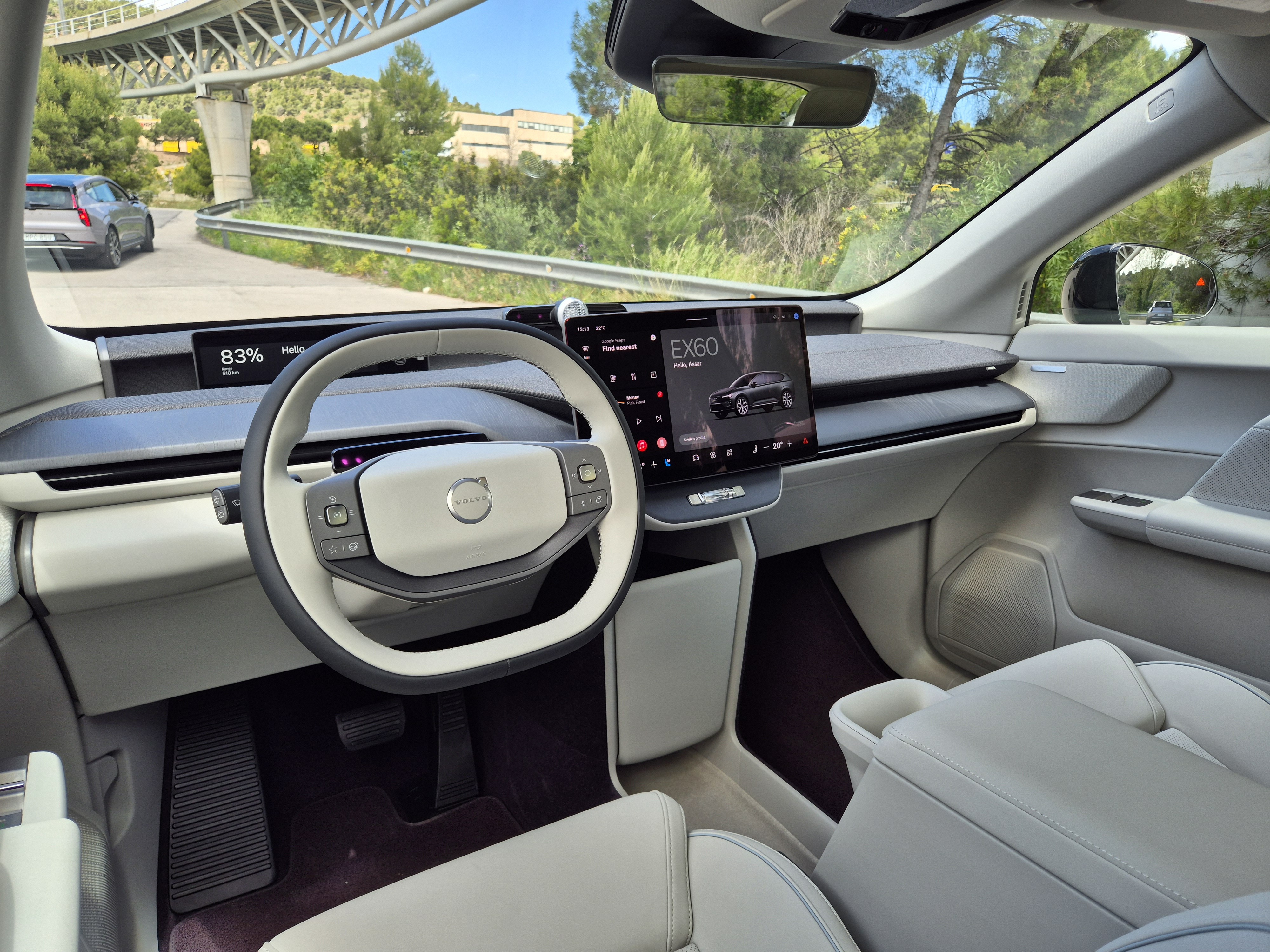 Interior de um Volvo EX60 com volante branco e cinza, painel minimalista e tela central exibindo o modelo. Ao fundo, uma estrada sinuosa com ponte e vegetação verde sob céu azul