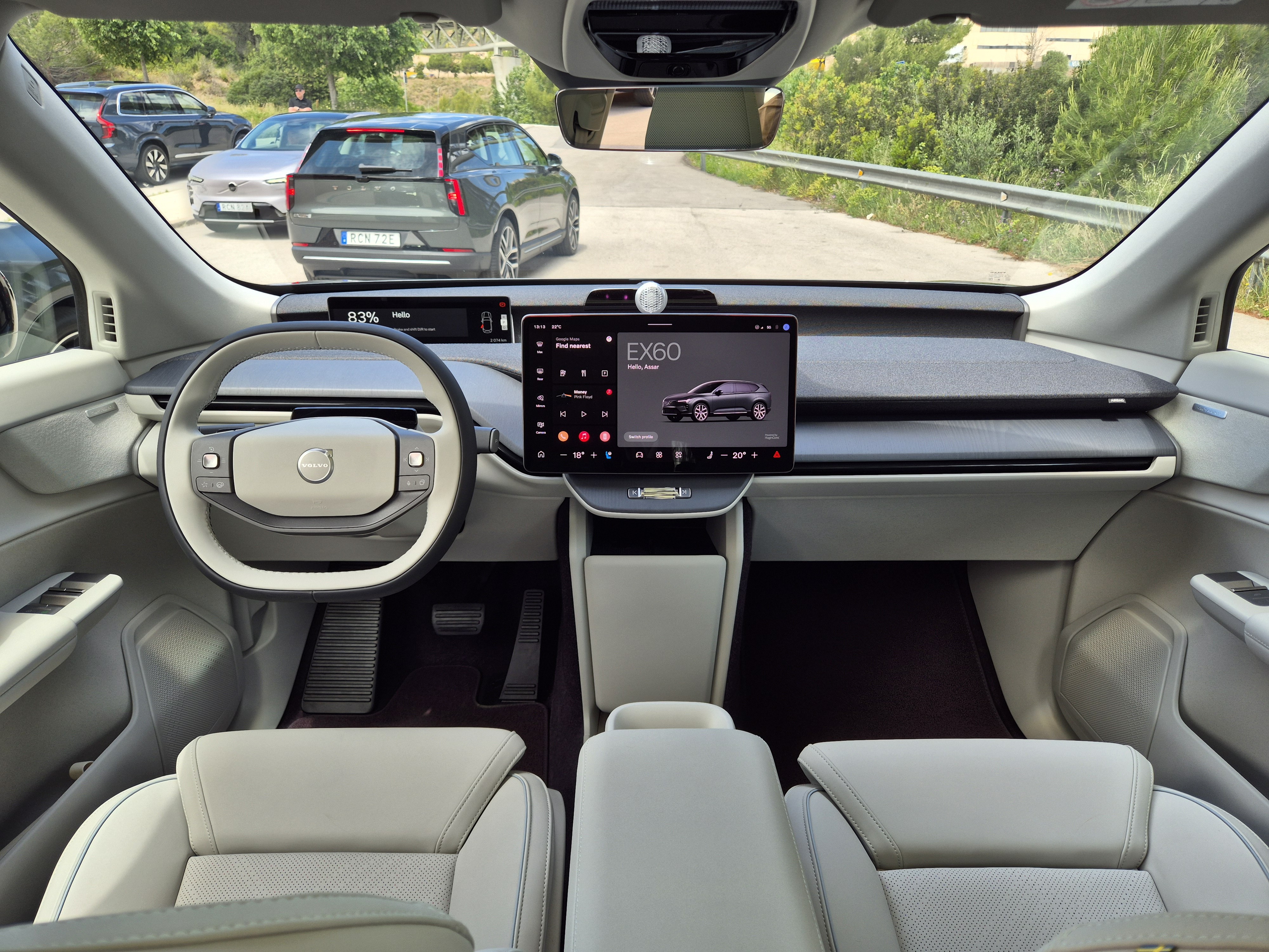 Interior de um carro Volvo EX60, com volante cinza e branco, tela central exibindo o modelo do veículo e carros estacionados ao fundo em uma estrada