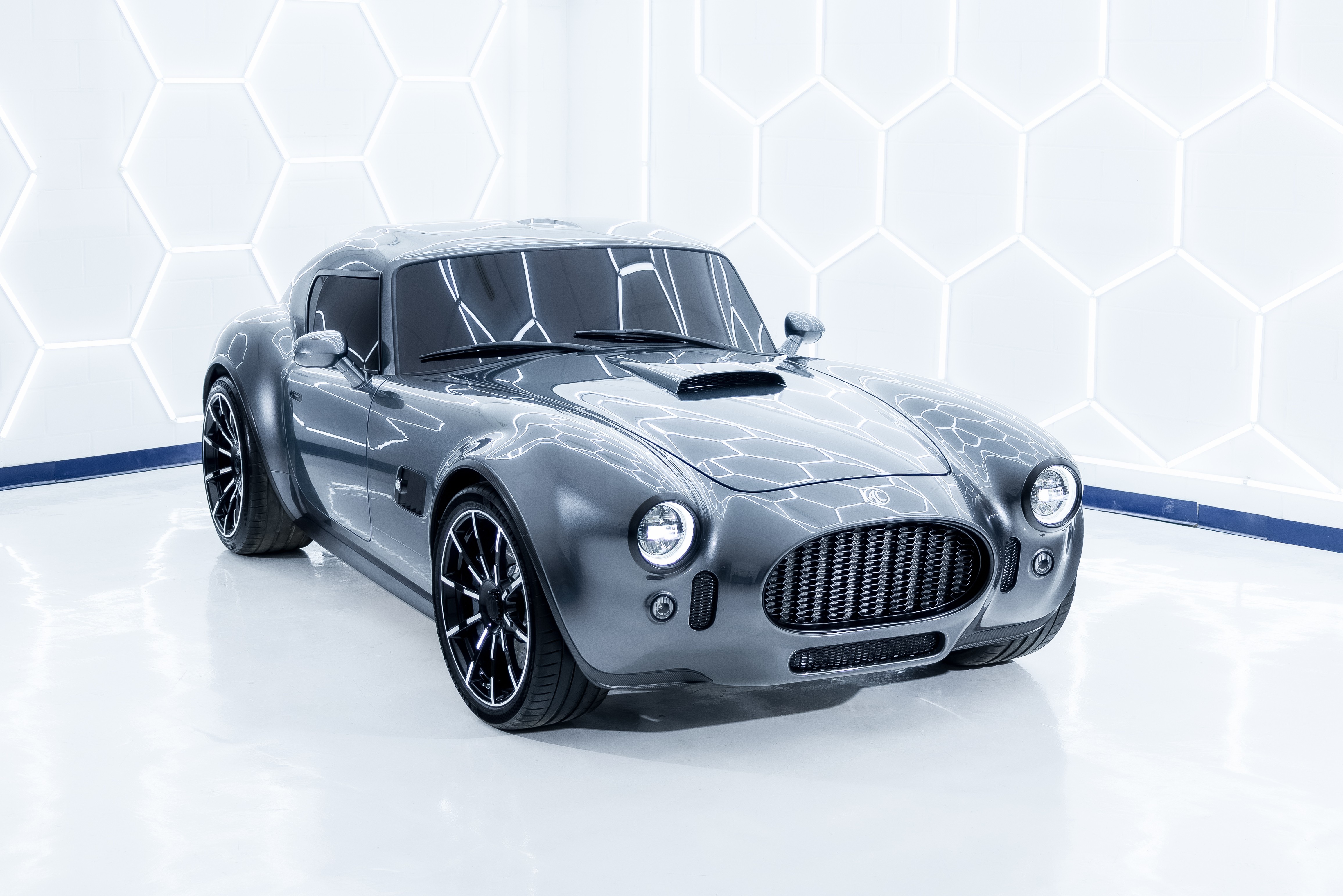 Carro esportivo cinza escuro, estilo clássico, com capô longo e entrada de ar, rodas pretas e detalhes cromados, em um estúdio branco com iluminação hexagonal na parede