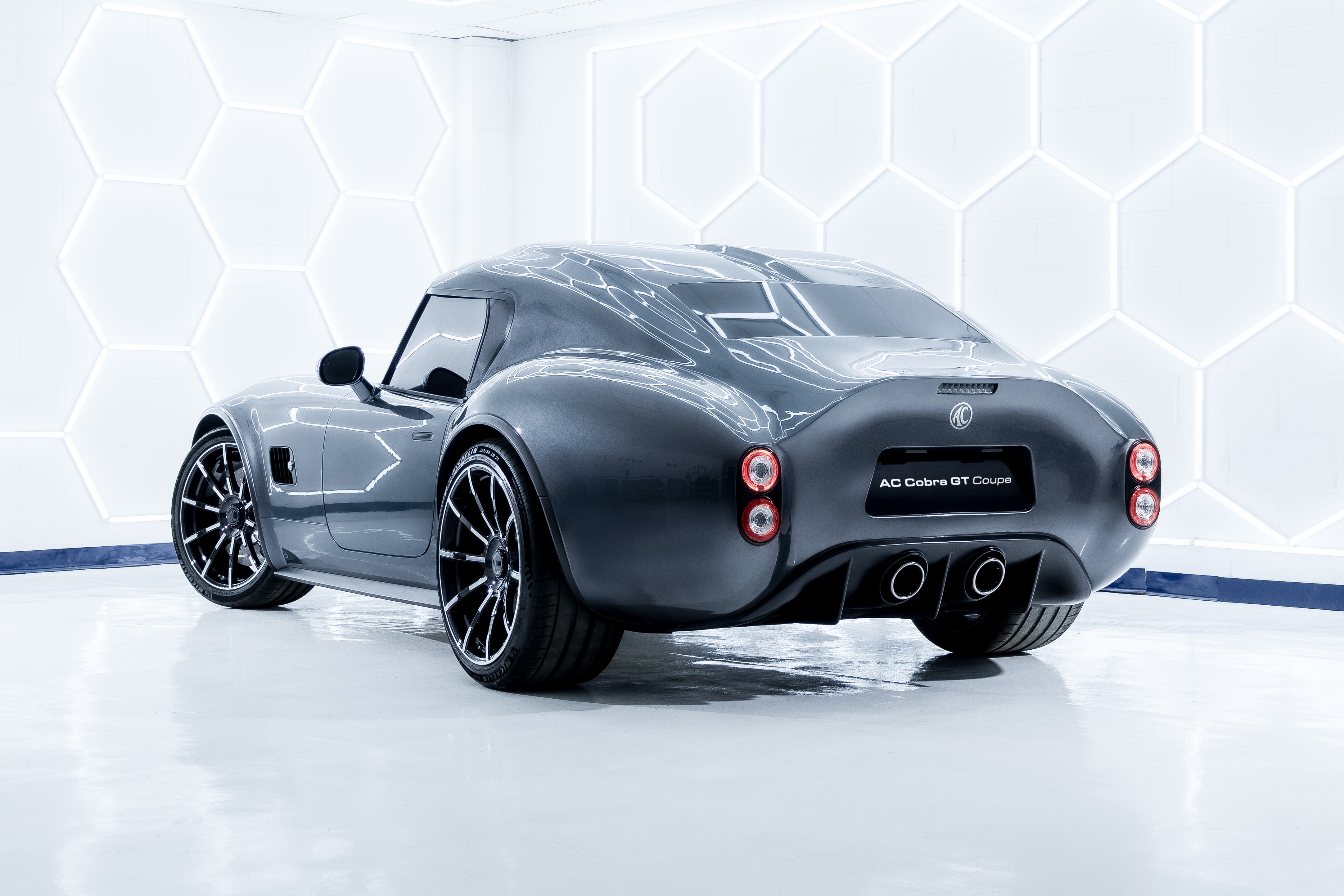 Carro esportivo AC Cobra GT Coupe cinza escuro, visto por trás, com rodas pretas e detalhes cromados, em um estúdio branco com iluminação hexagonal