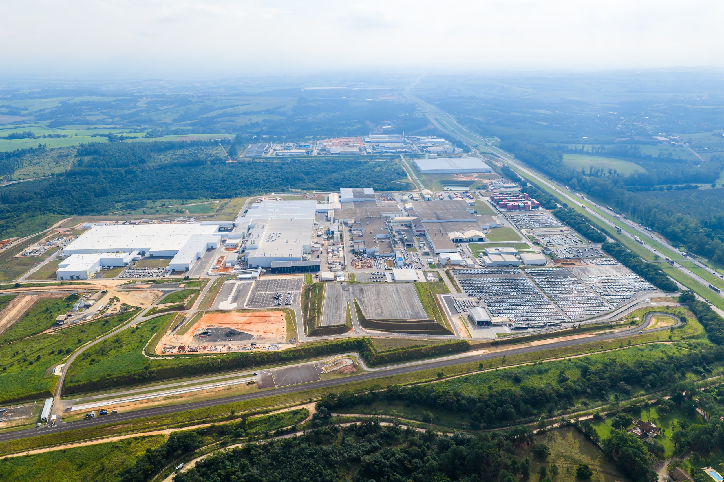 Vista aérea de uma grande fábrica automotiva com telhados brancos, cercada por vastos estacionamentos cheios de carros novos. Uma rodovia movimentada corre paralela à fábrica, e o cenário inclui áreas verdes, campos e outras construções industriais distantes sob um céu claro