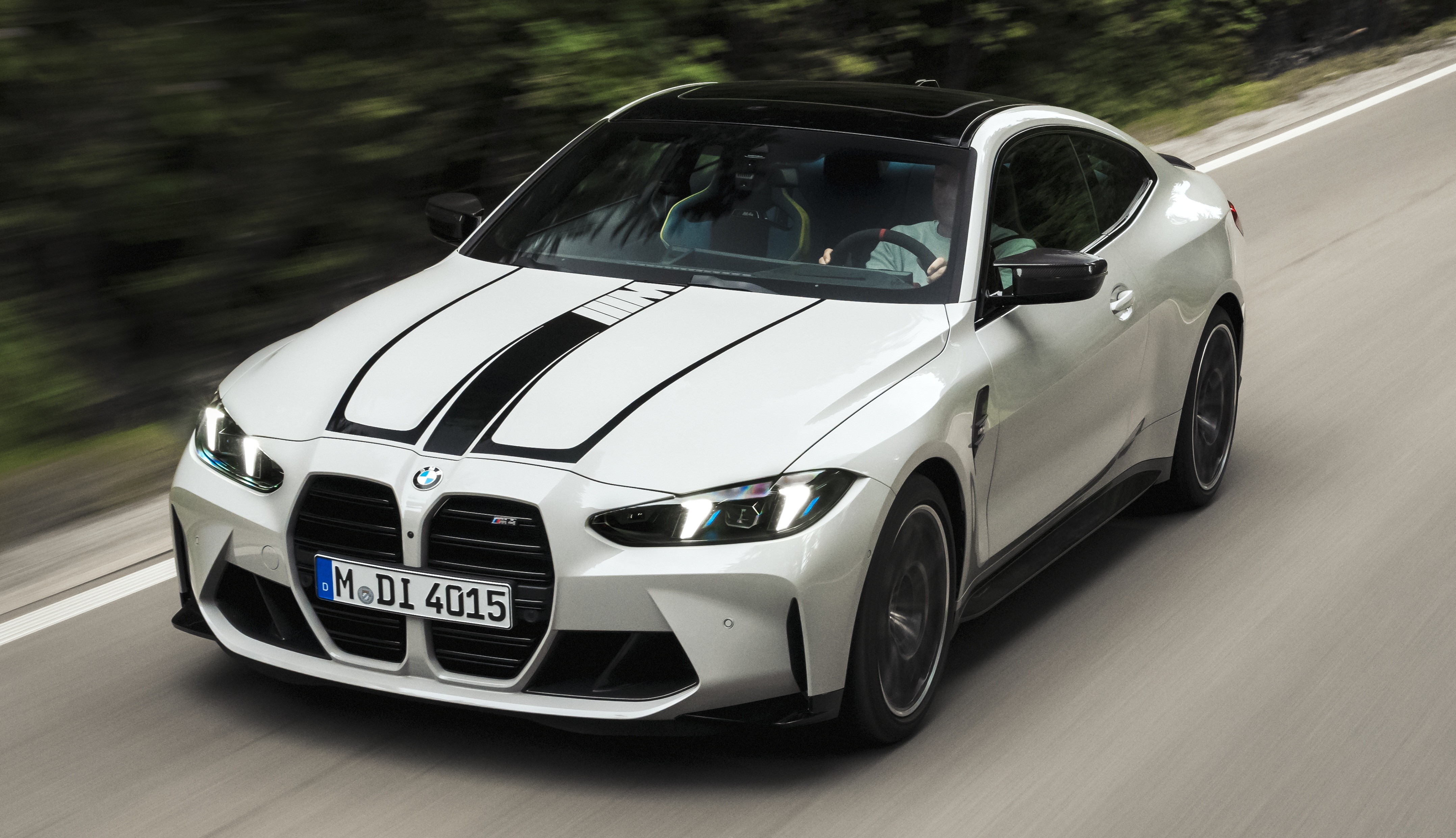 BMW M4 branco com teto preto e duas faixas pretas no capô, em movimento rápido numa estrada asfaltada com árvores borradas ao fundo