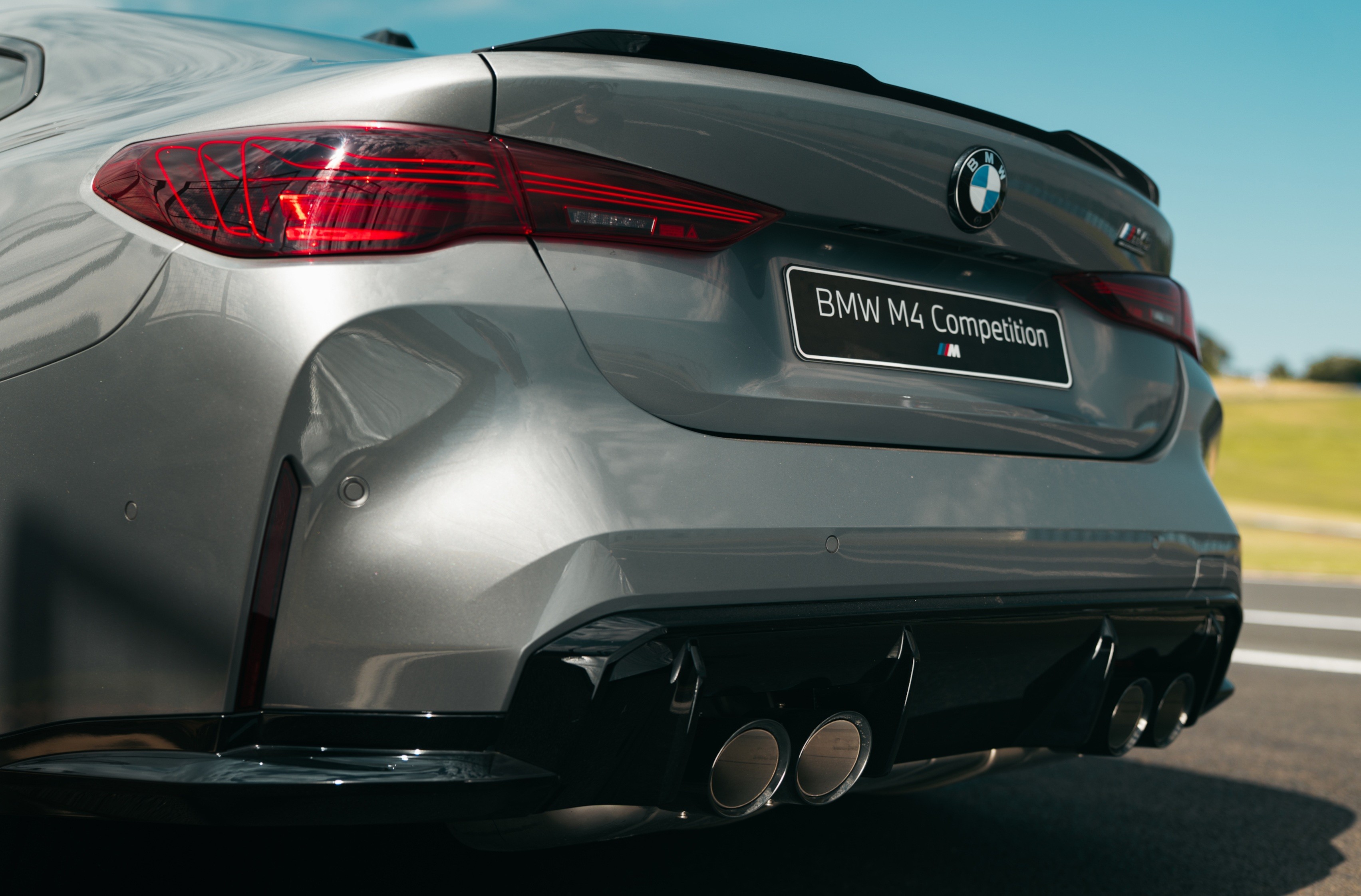 Traseira de um carro BMW M4 Competition cinza metálico, com lanternas vermelhas, emblema da marca e quatro saídas de escapamento cromadas, em uma pista de corrida sob céu azul