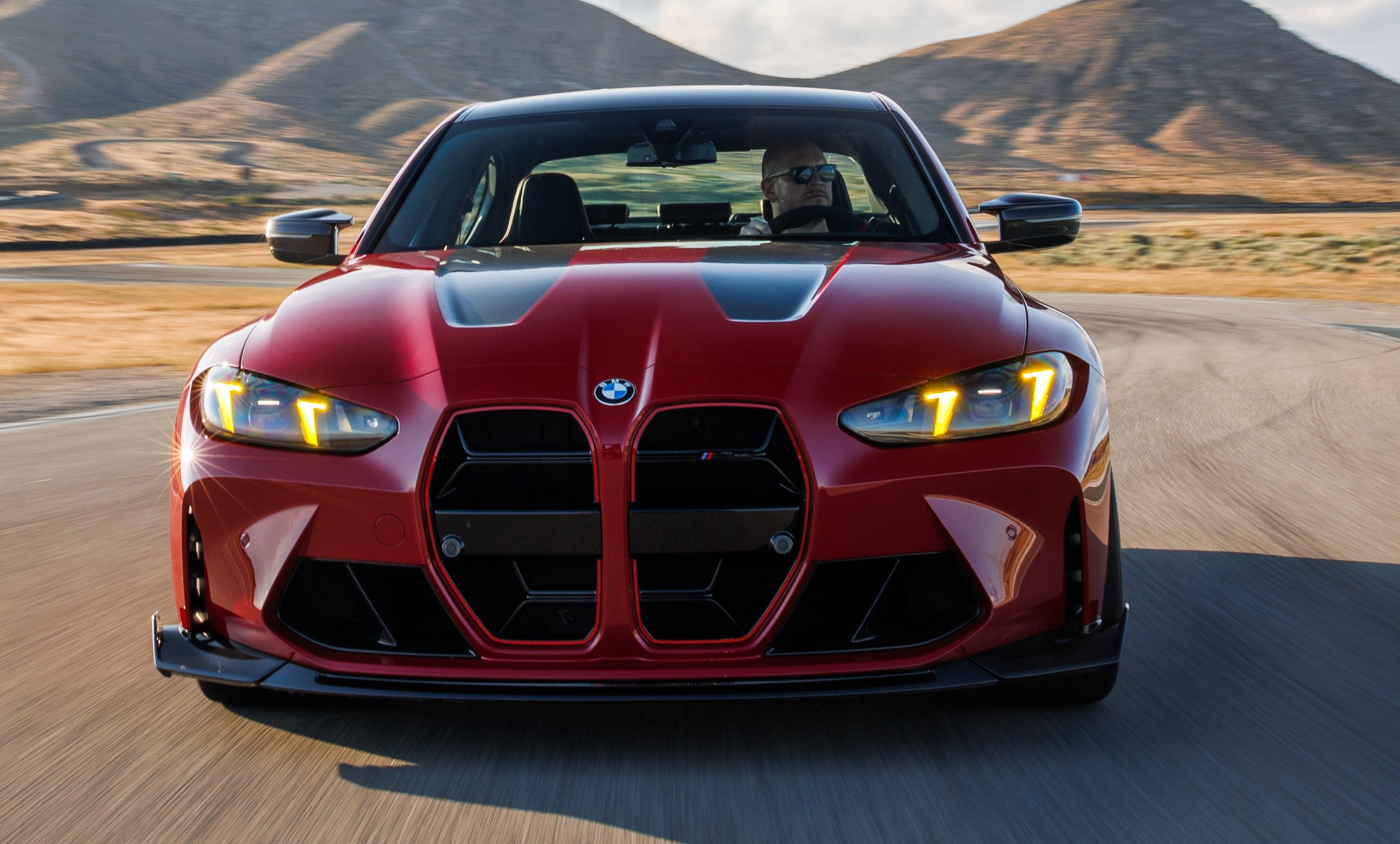Carro esportivo vermelho BMW M4 Competition, com motorista usando óculos escuros, em alta velocidade numa pista sinuosa com montanhas ao fundo