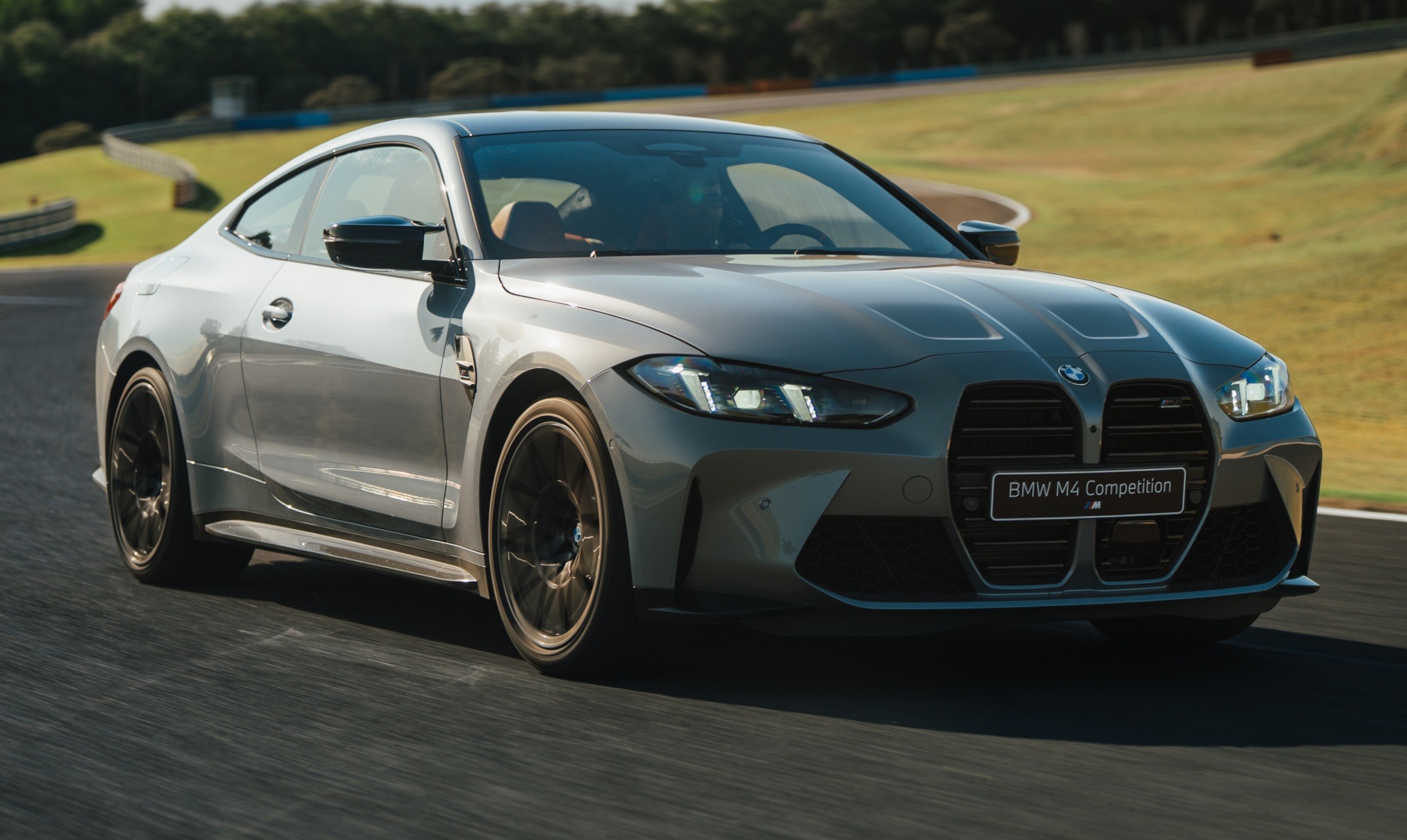 Carro BMW M4 Competition cinza met&aacute;lico em movimento numa pista de corrida, com gramado verde e &aacute;rvores ao fundo