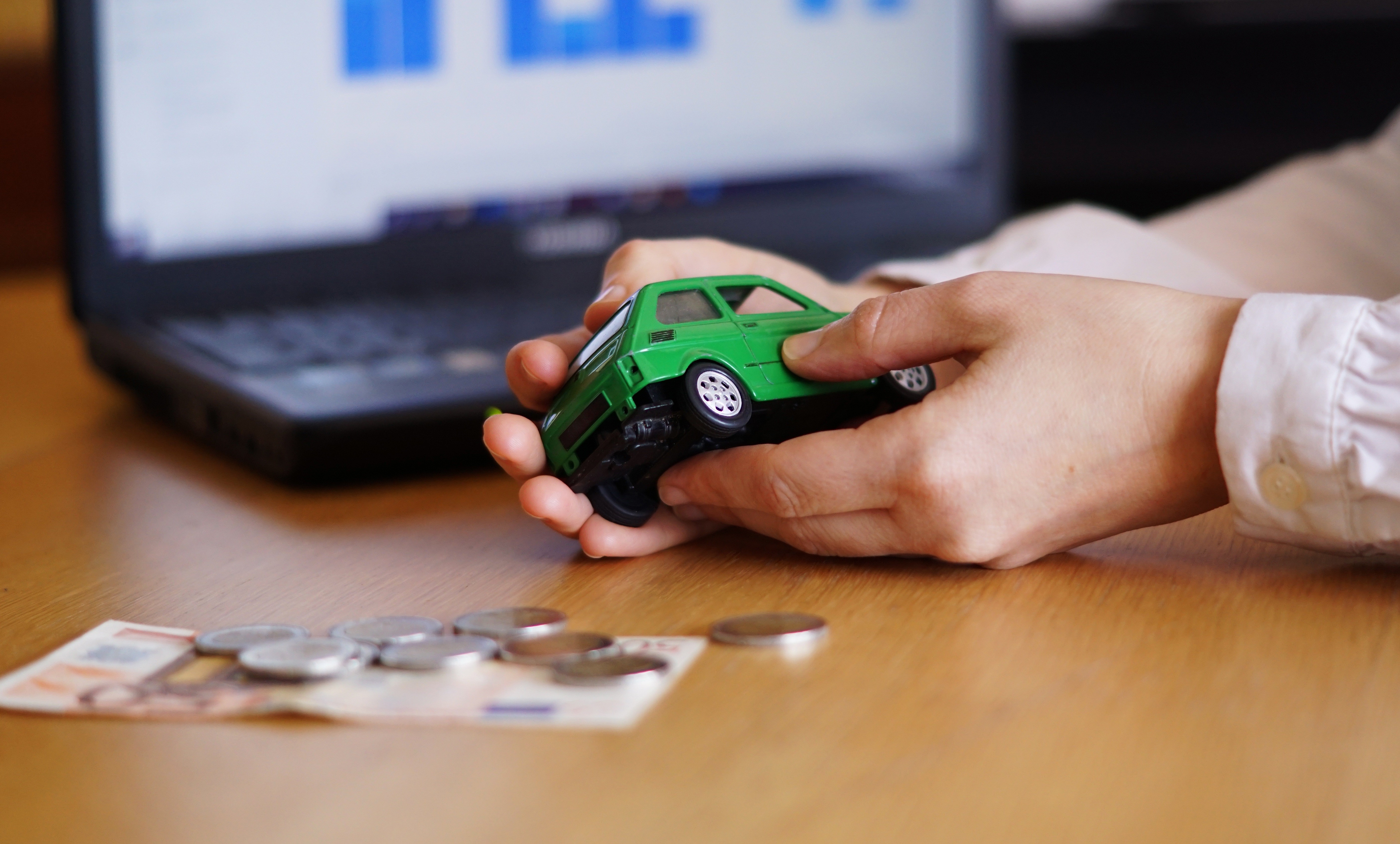 Mãos segurando um carro de brinquedo verde sobre uma mesa de madeira, com notas de euro e moedas espalhadas, e um notebook com gráficos ao fundo