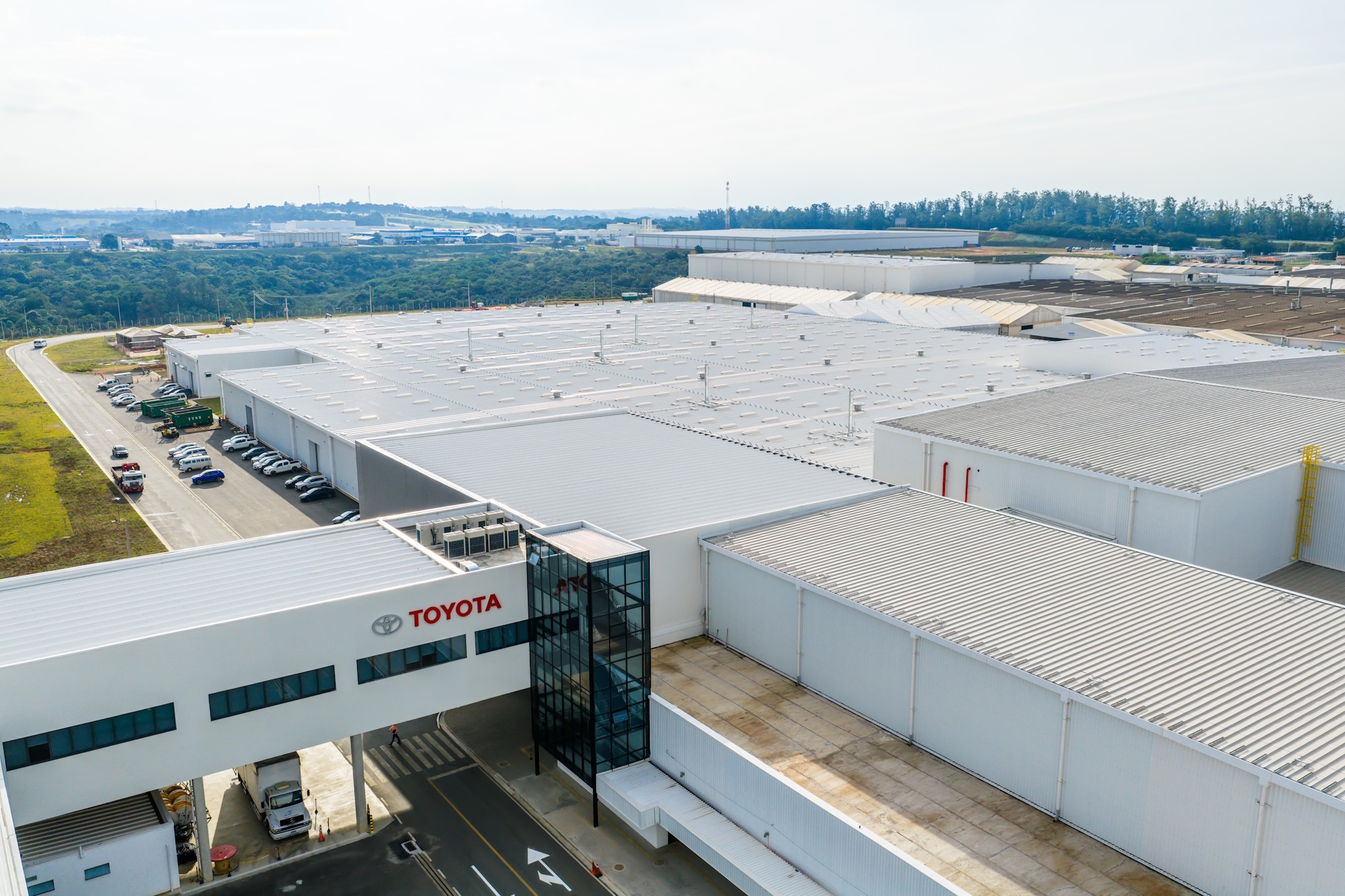 Vista aérea de uma grande fábrica da Toyota com telhados brancos e extensos, conectada por uma passarela elevada com o logotipo da empresa. Carros e caminhões estacionados em pátios, e uma área verde com árvores ao fundo, sob um céu claro