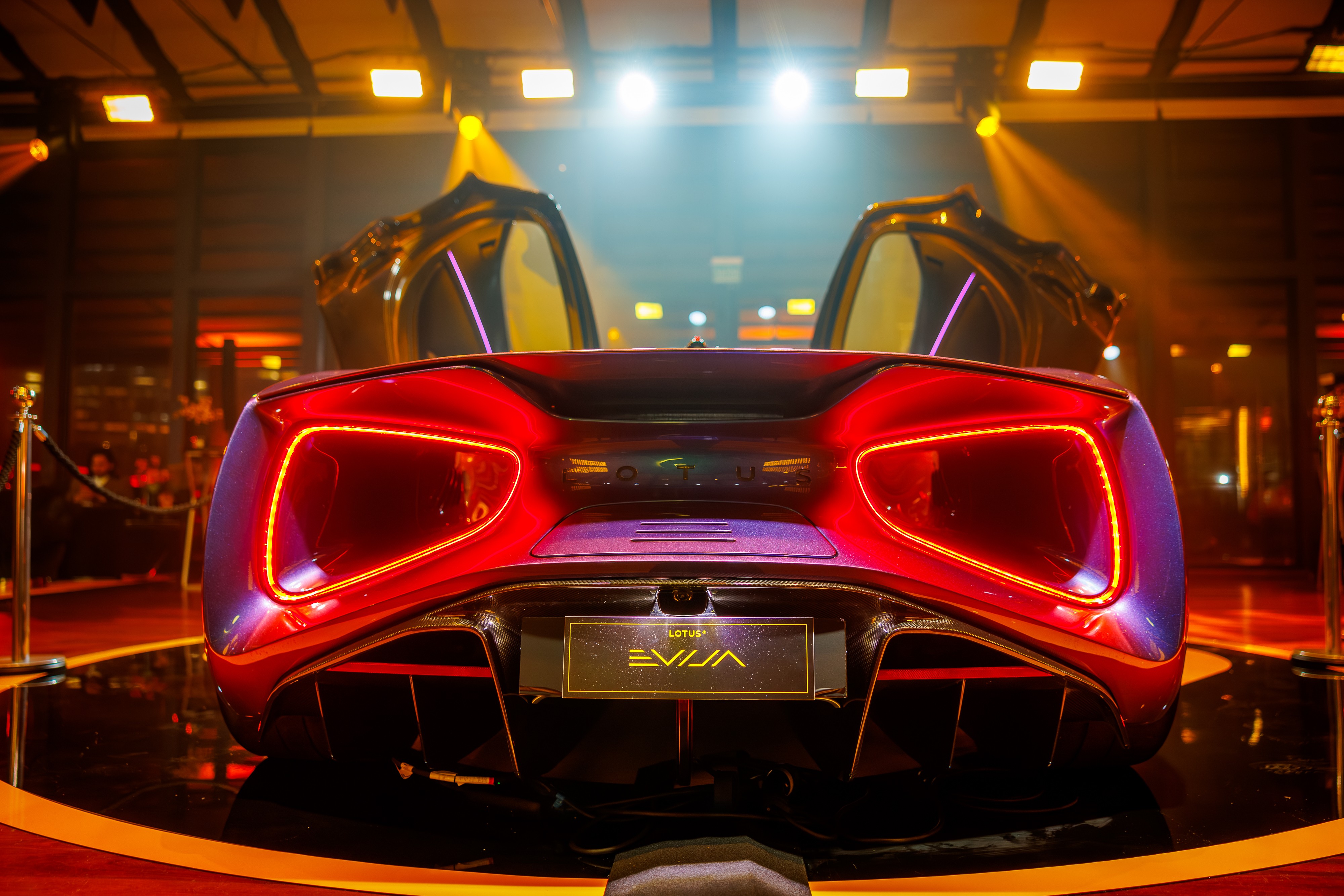 Traseira de um carro esportivo vermelho brilhante, modelo Lotus Evija, com as portas abertas para cima, exibindo luzes traseiras em neon e um difusor aerodinâmico. O veículo está em um palco escuro com iluminação laranja e roxa, cercado por pilares dourados e cordões de veludo, sugerindo um evento de lançamento ou exposição