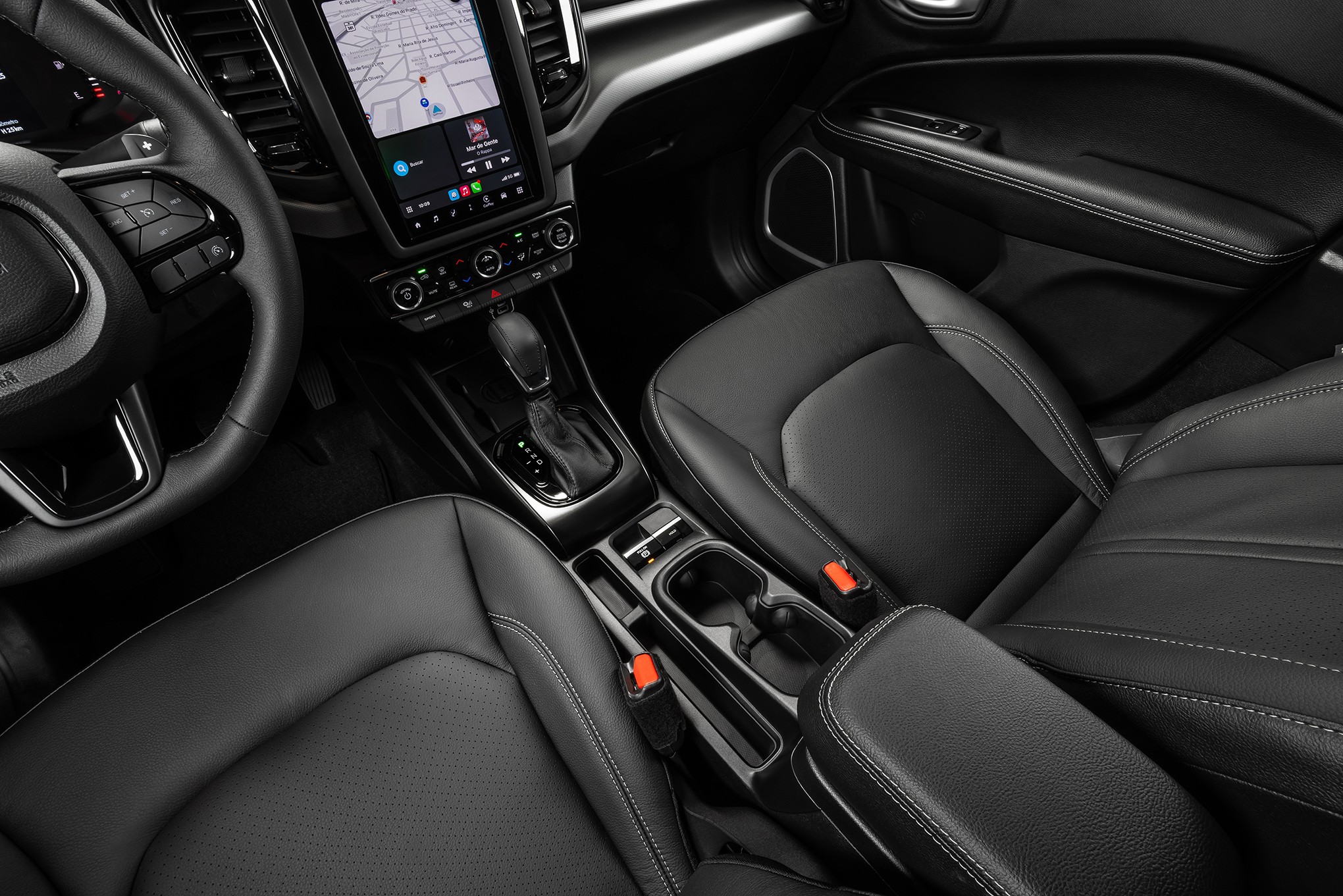 Interior de carro moderno com bancos de couro preto, console central com porta-copos, câmbio automático e tela multimídia exibindo mapa de navegação. Volante multifuncional à esquerda