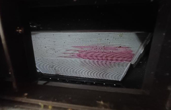 Radiador de carro com aletas metálicas horizontais e mancha rosa avermelhada de vazamento de fluido, em um compartimento escuro