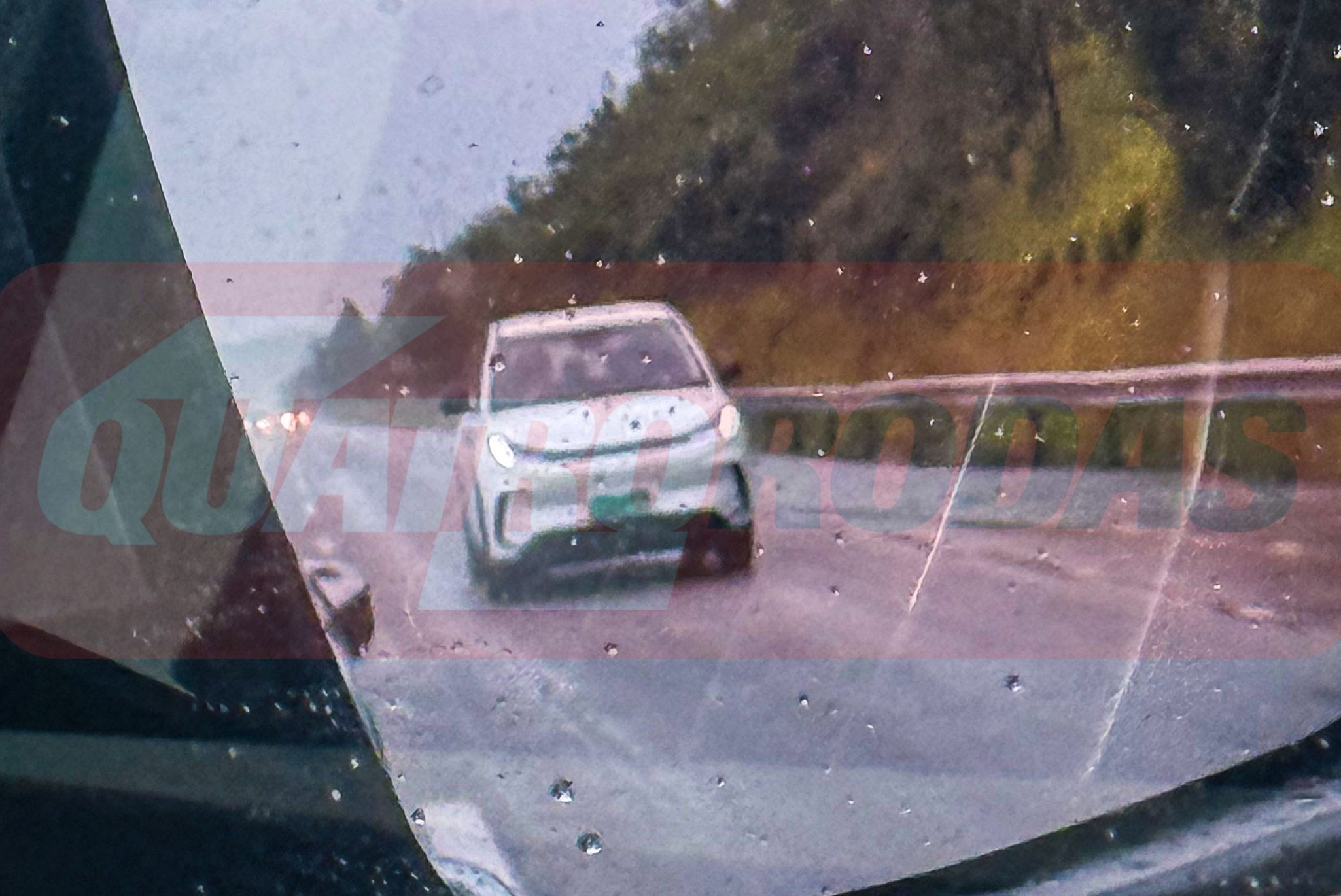 Espelho retrovisor lateral de um carro, molhado pela chuva, refletindo um veículo branco na estrada sob um céu nublado e montanhas ao fundo
