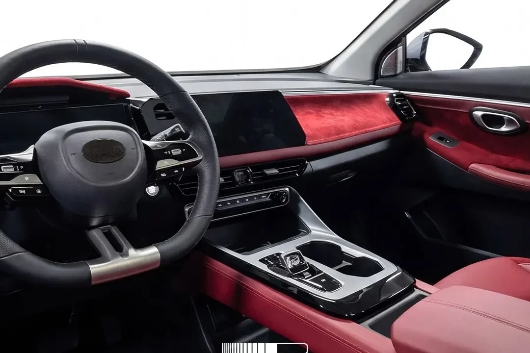 Interior de carro moderno com volante preto, painel em veludo vermelho e tela digital ampla. Detalhes em preto brilhante e bancos de couro vermelho. O console central possui porta-copos e controles de marcha. O retrovisor interno é visível no topo.