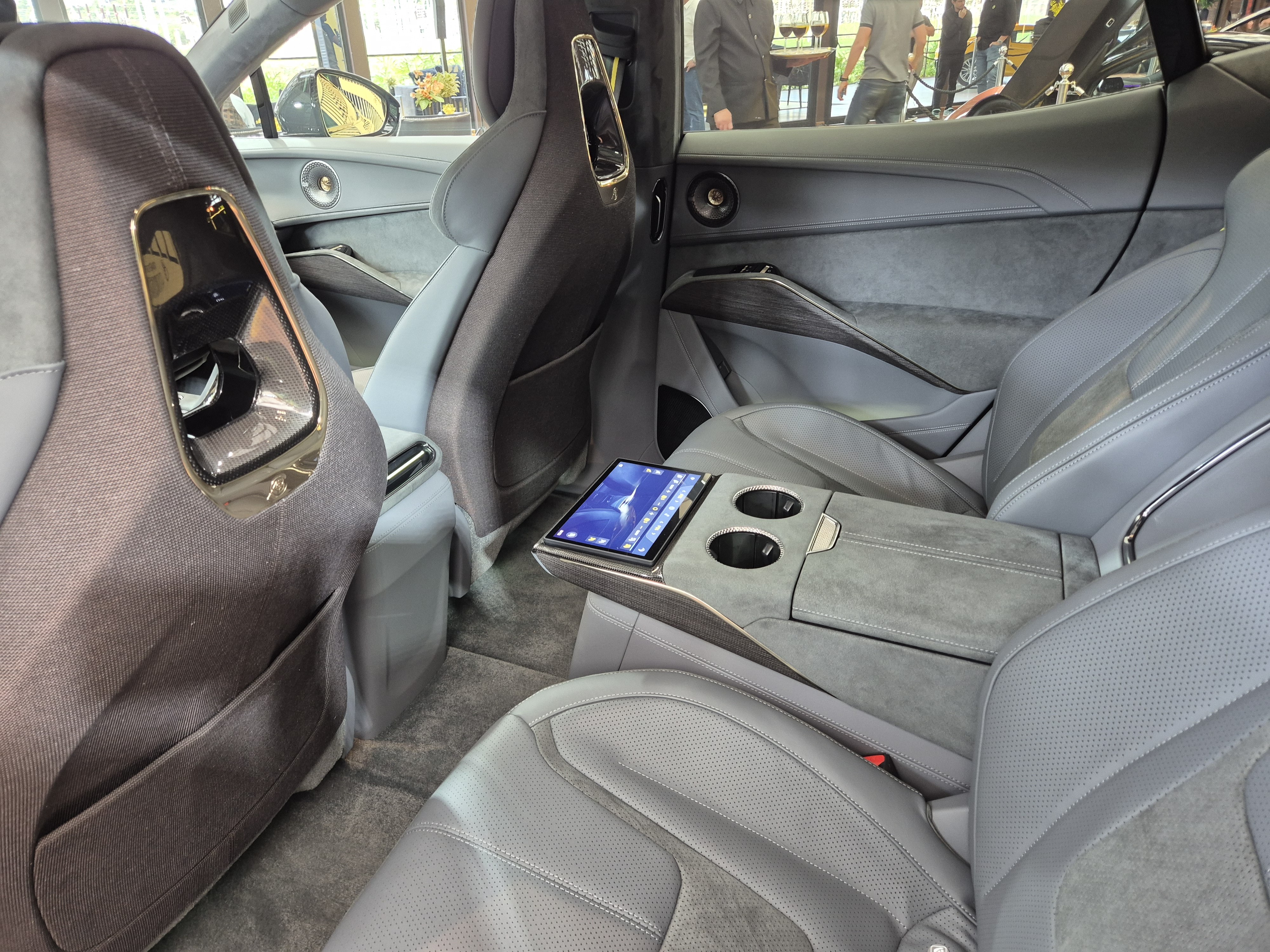 Interior de carro luxuoso com bancos de couro cinza e detalhes em camurça. Um tablet com tela azul está no console central traseiro, ao lado de dois porta-copos. Pessoas desfocadas aparecem ao fundo, fora do veículo