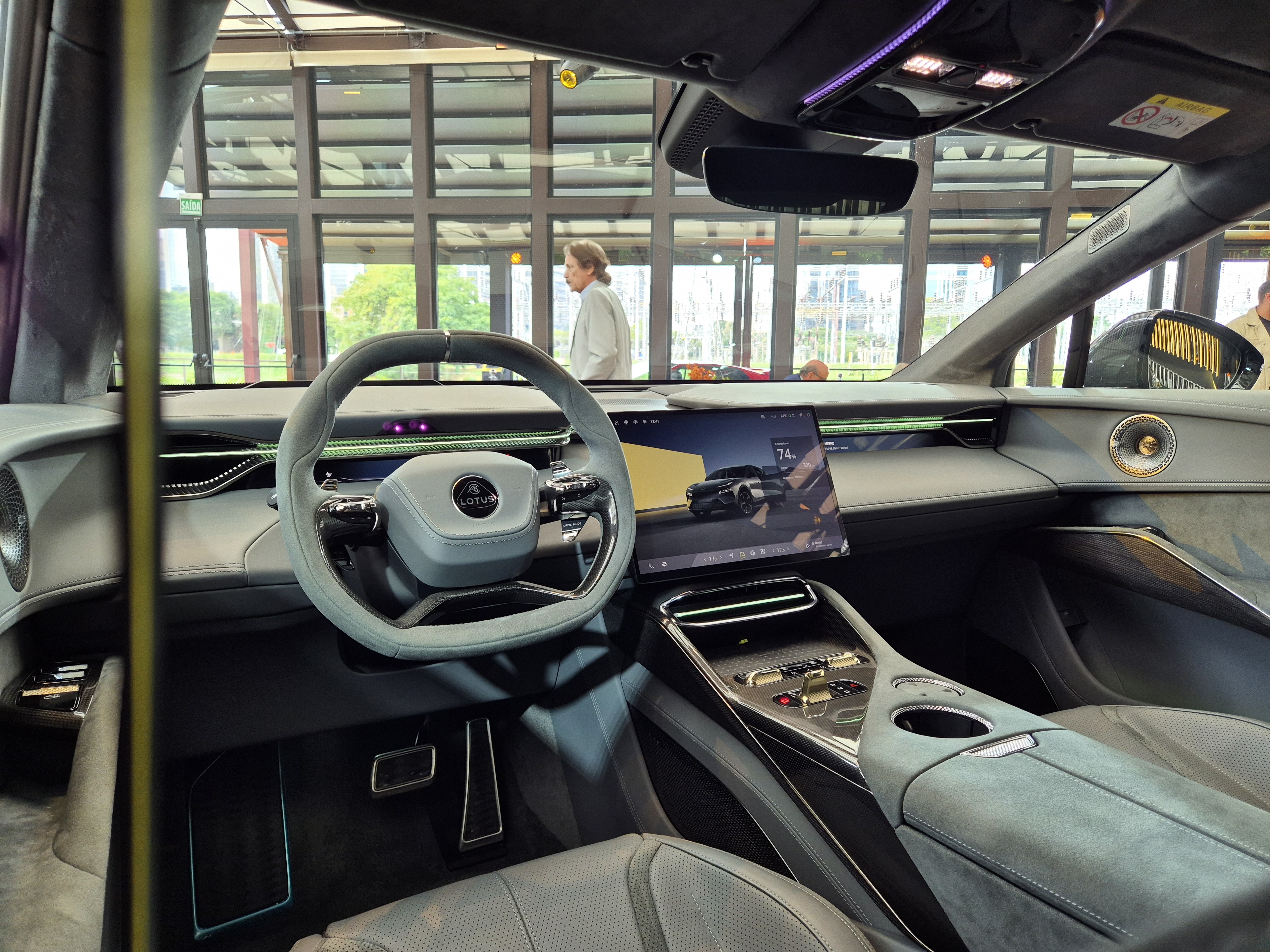 Interior de um carro Lotus cinza com volante esportivo, tela multimídia exibindo outro carro e console central com botões e porta-copos. Uma pessoa de camisa clara caminha ao fundo, visível através do para-brisa.