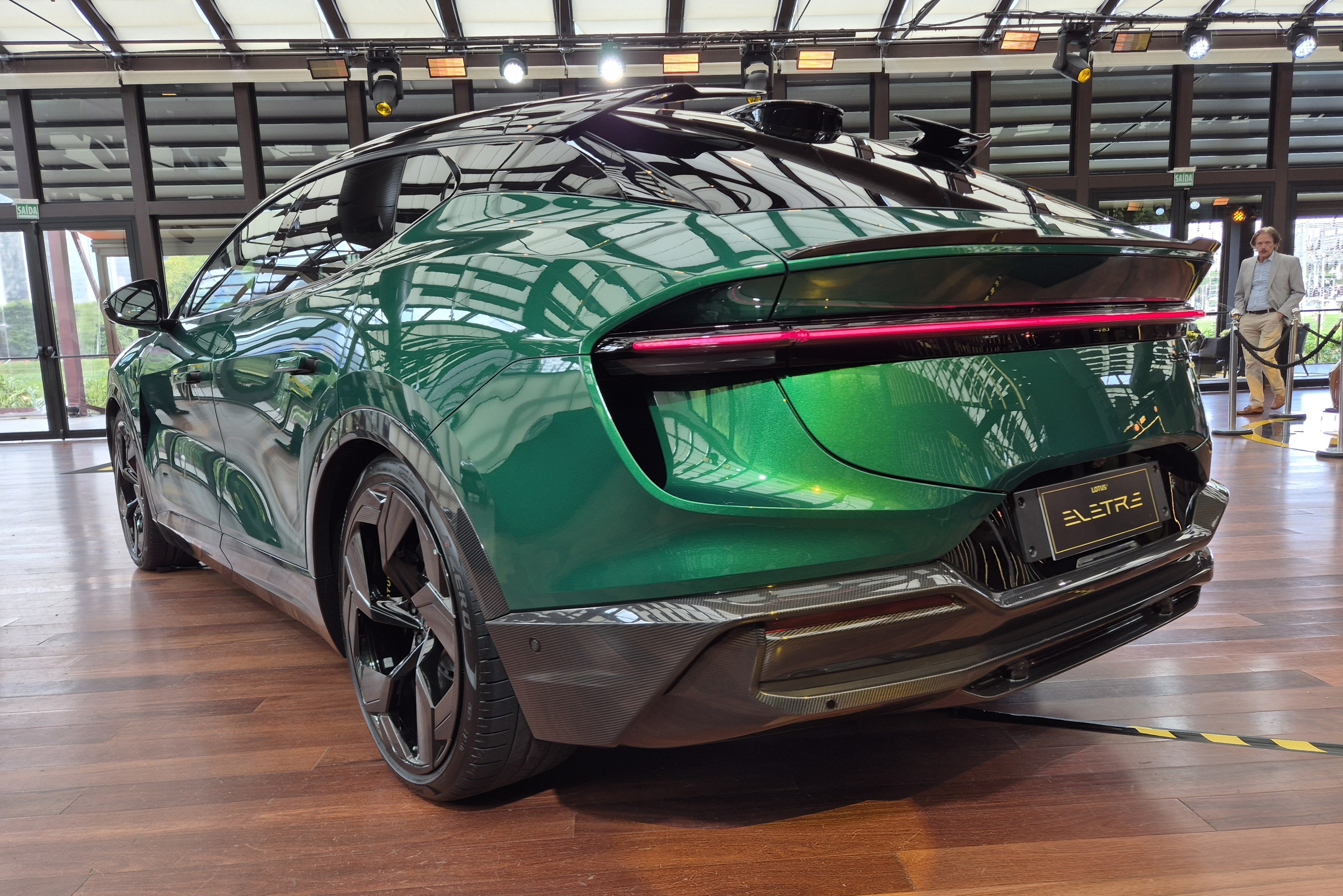 Traseira de um carro esportivo verde metálico, modelo Lotus Eletre, com lanterna vermelha horizontal e aerofólio duplo. O veículo está em um piso de madeira, com um homem de blazer claro ao fundo, próximo a uma janela.