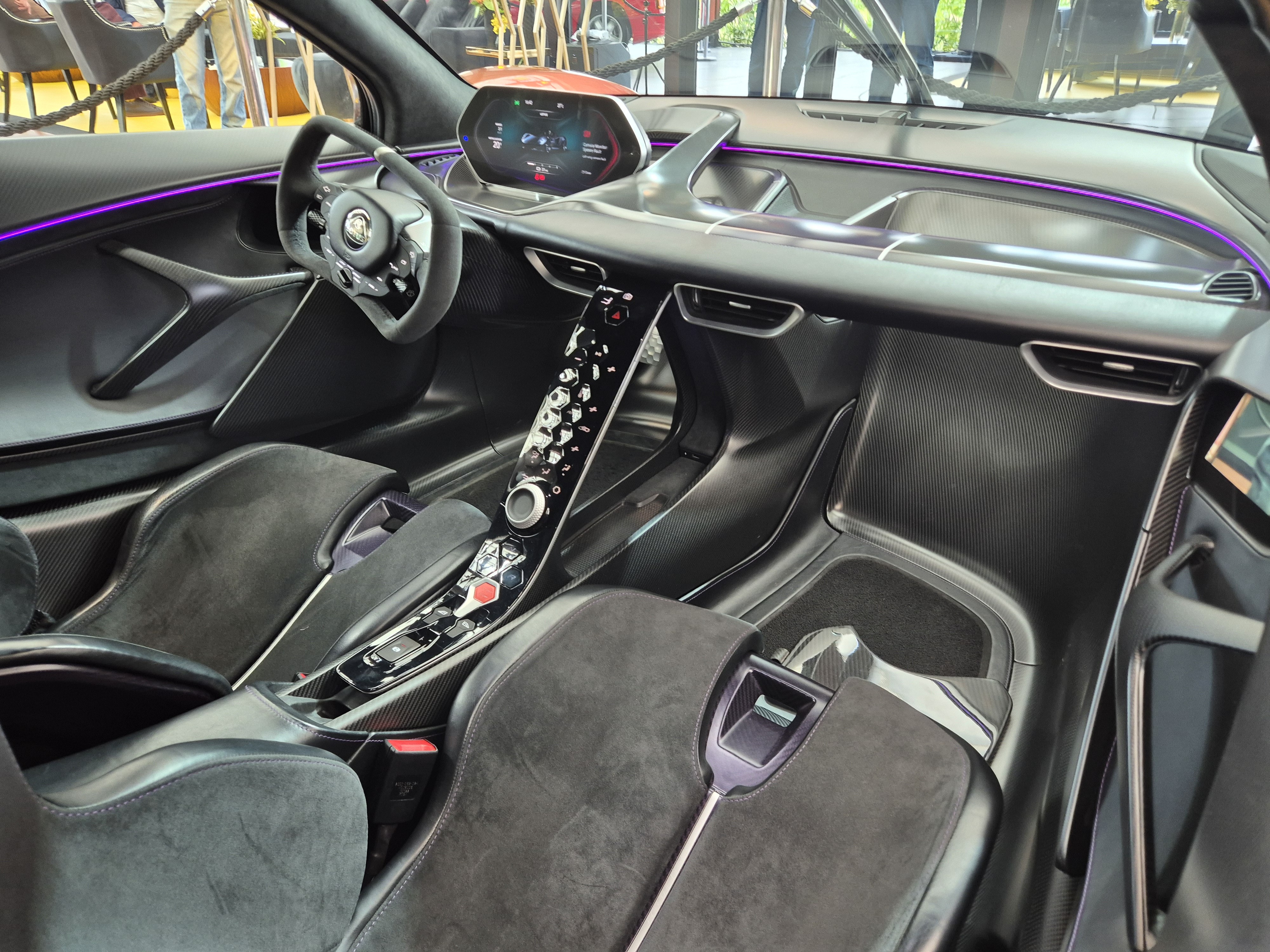 Interior de um carro esportivo moderno, com bancos de camurça preta, volante esportivo, painel digital e console central com botões futuristas. Uma linha de luz ambiente roxa percorre o painel, destacando o design arrojado.