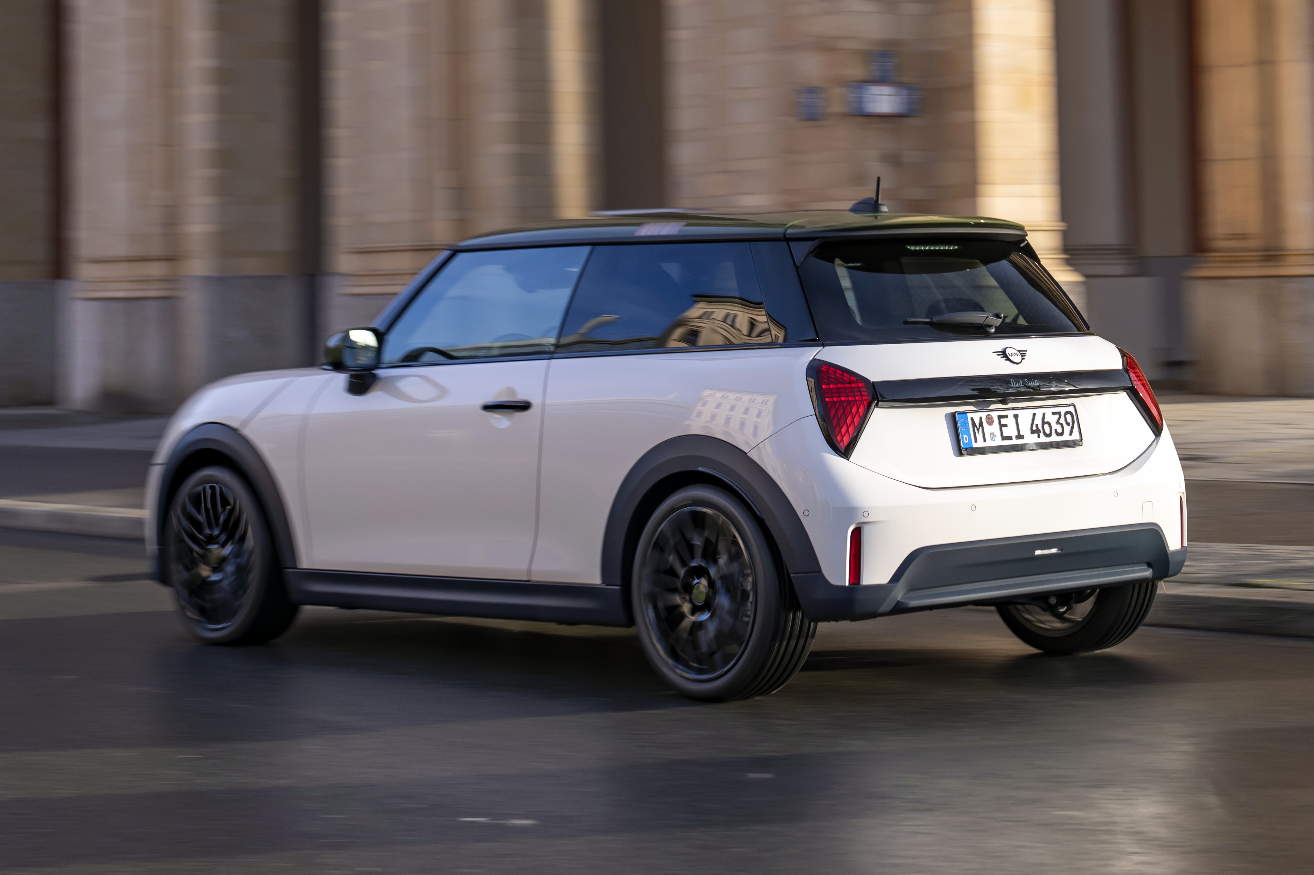 Mini Cooper branco com teto preto e rodas escuras, visto de traseira lateral, em movimento rápido sobre asfalto, com fundo de prédio borrado