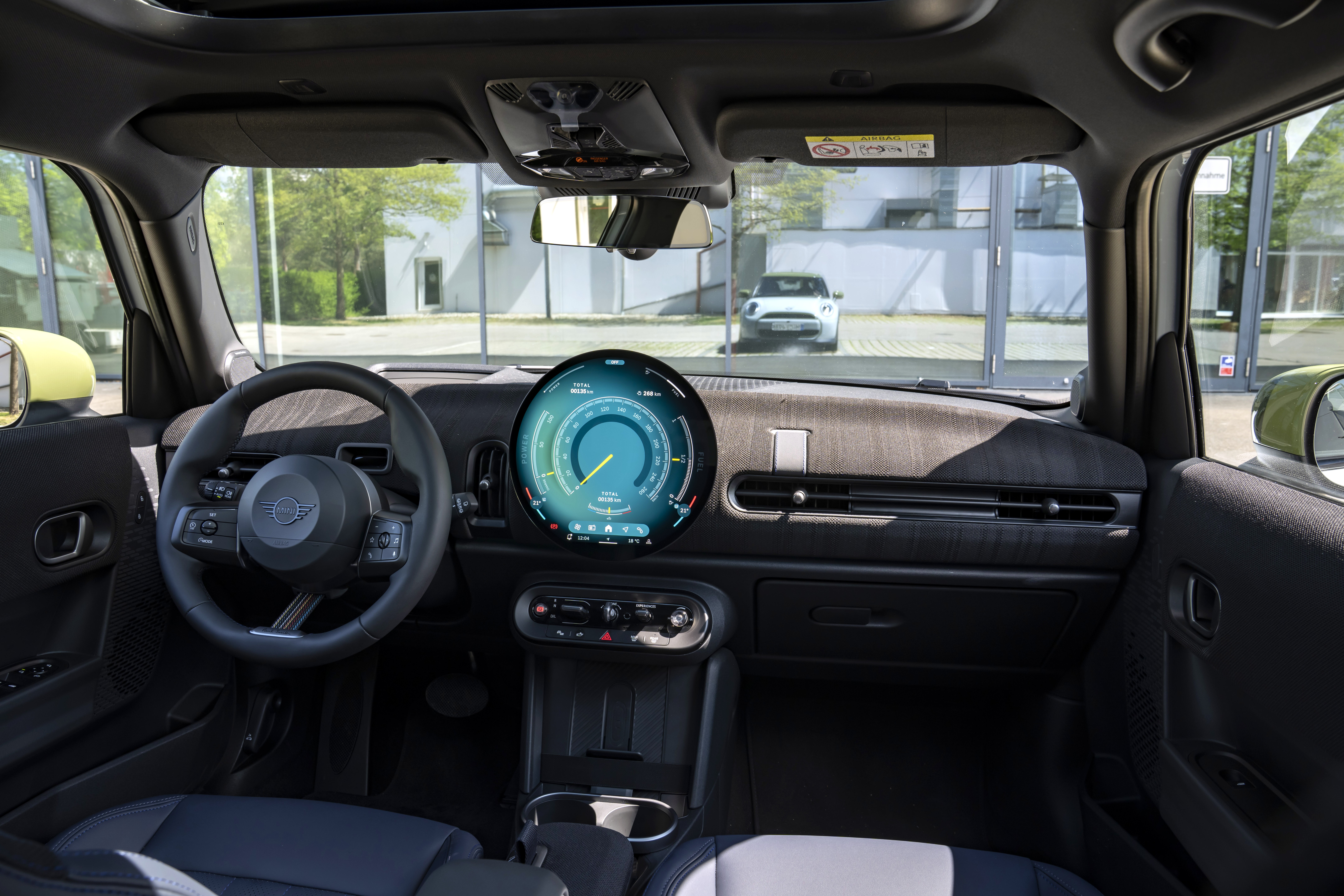 Interior de um carro Mini Cooper, com volante à esquerda e um painel digital redondo exibindo informações do veículo. Ao fundo, um carro branco estacionado em frente a um prédio com grandes janelas de vidro