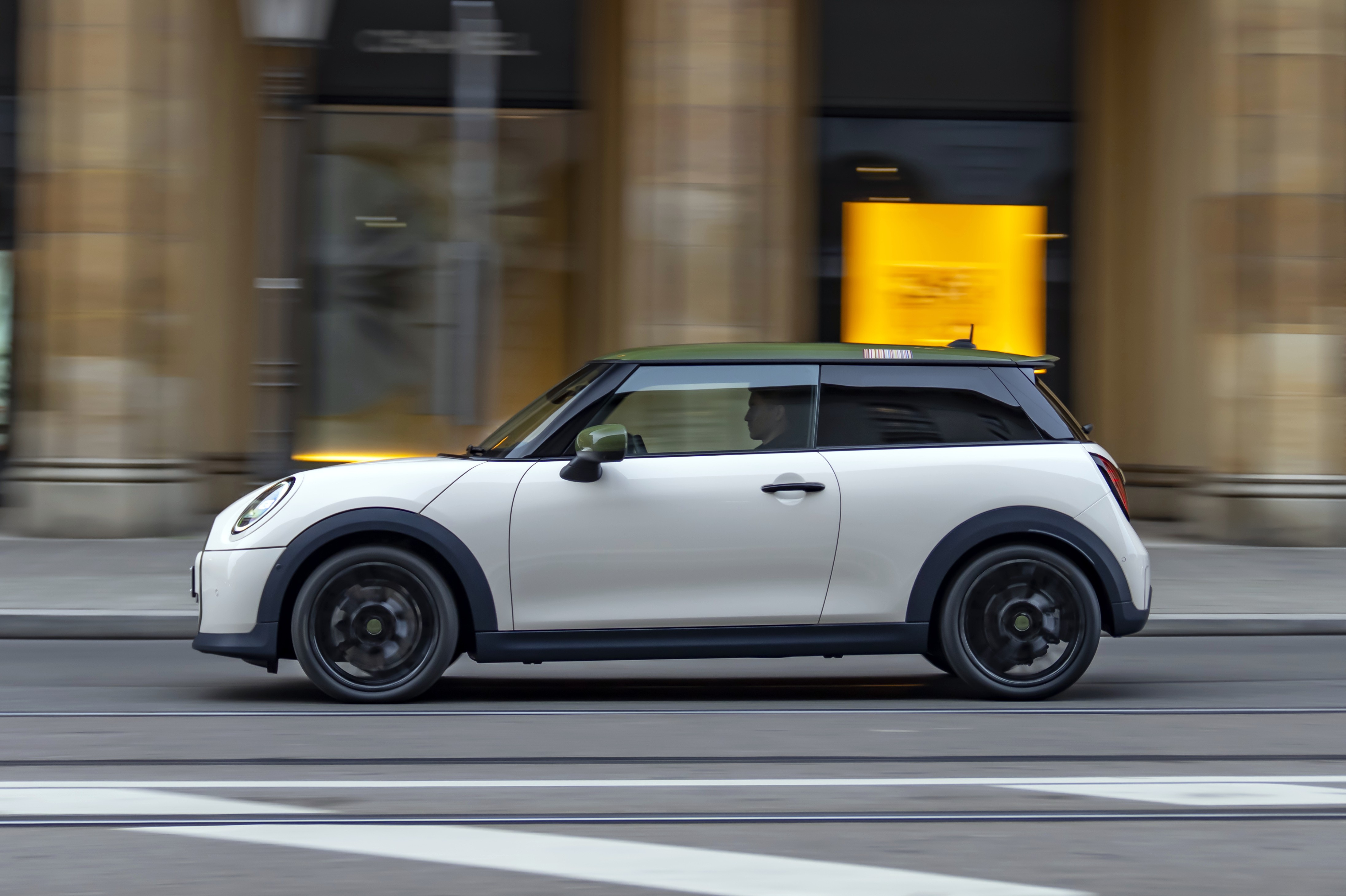 Mini Cooper branco com teto verde-oliva e rodas pretas em movimento, visto de lado, com um motorista visível. O fundo desfocado sugere velocidade, com edifícios de fachada clara e uma placa amarela. Há faixas brancas no asfalto em primeiro plano