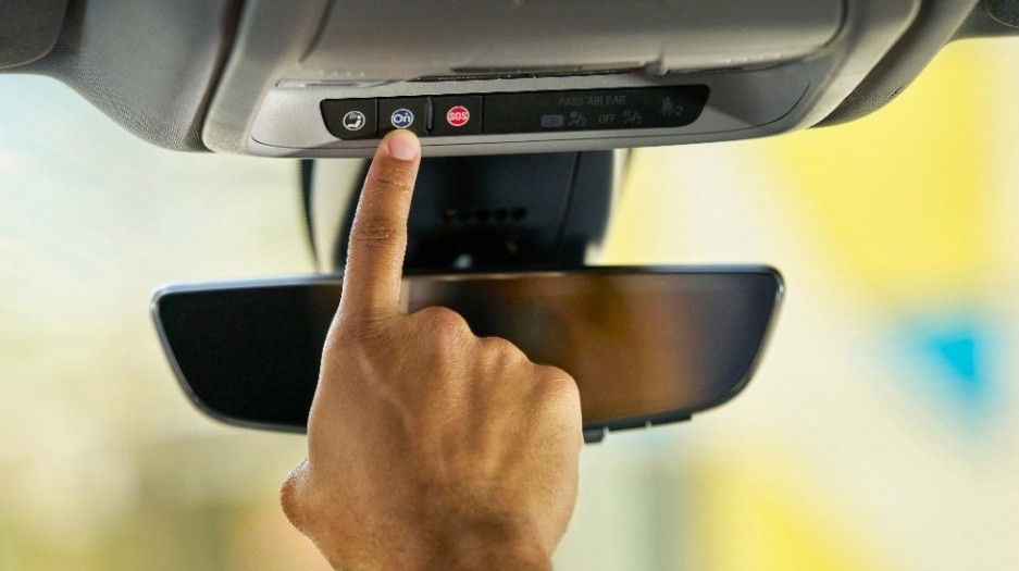 Dedo indicador de uma pessoa pressionando o botão azul de um sistema de assistência veicular, localizado no console superior do carro, acima do retrovisor interno. Há também um botão vermelho de SOS e outro com um ícone de telefone