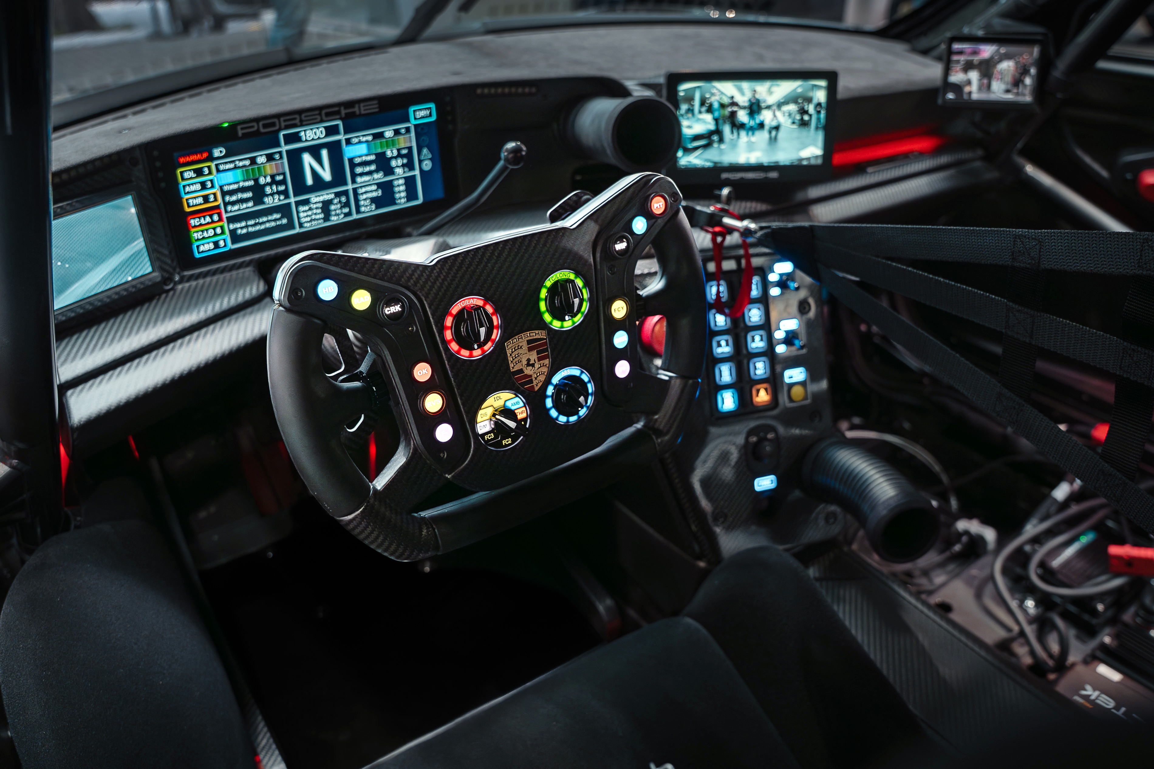 Interior de um carro de corrida com volante multifuncional preto e botões coloridos iluminados. Painel digital exibe informações do veículo e uma tela mostra pessoas em um ambiente urbano. O banco do motorista é preto e o cinto de segurança de quatro pontos está visível. Fios e componentes eletrônicos expostos no console central.