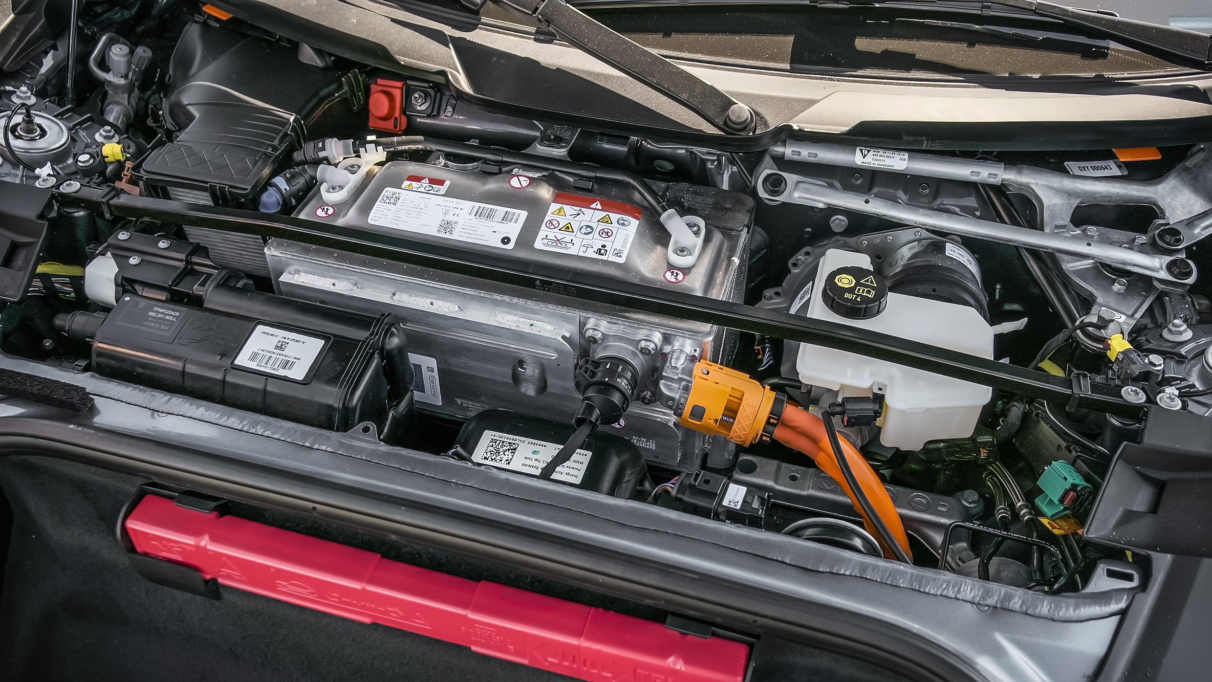 Compartimento do motor de um carro elétrico, com bateria prateada, cabos laranja e reservatório de fluido branco, além de um triângulo de segurança vermelho no porta-malas