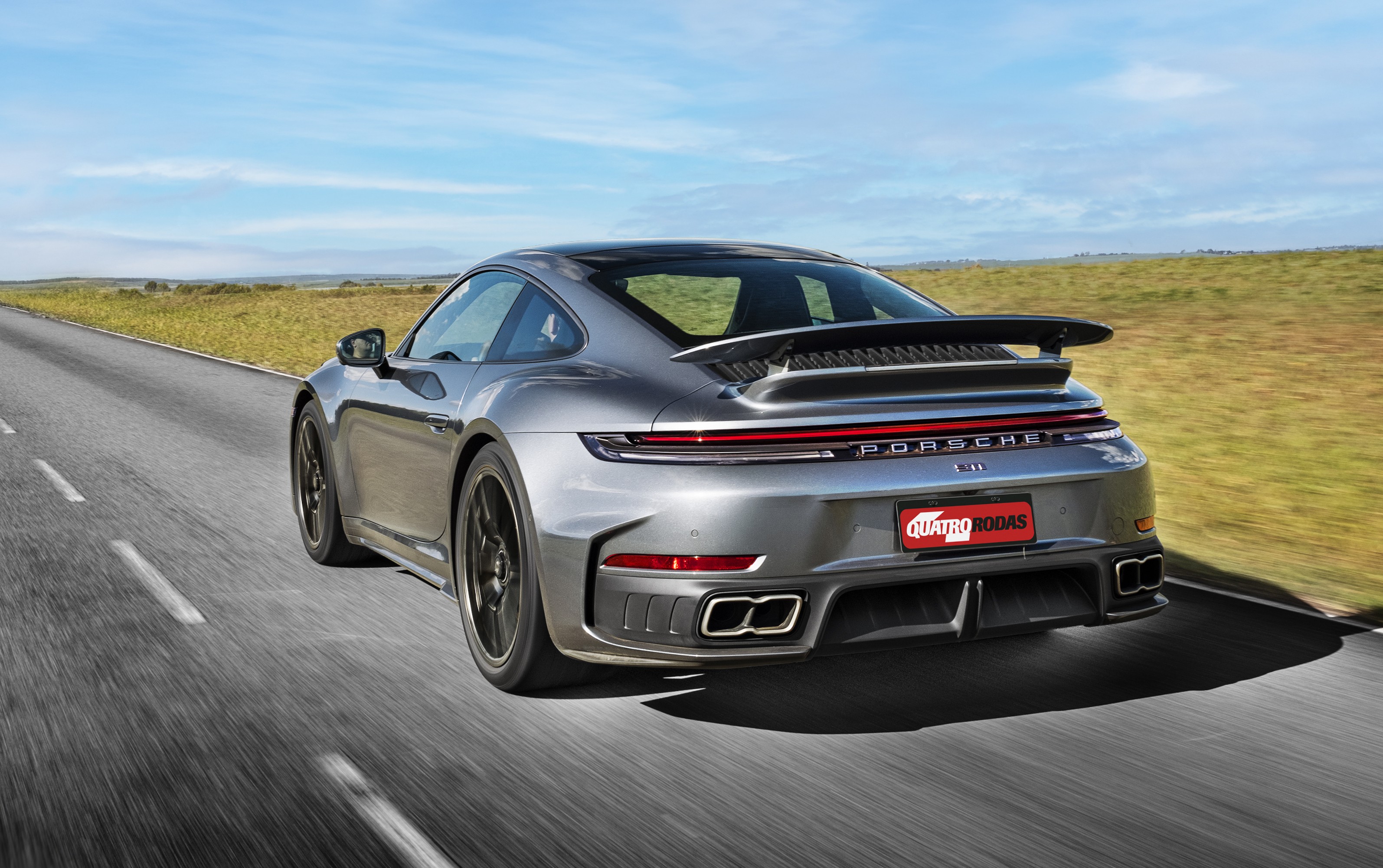 Porsche 911 cinza escuro em movimento, visto por trás, com aerofólio e escapamento duplo, em estrada asfaltada com vegetação ao lado, sob céu azul claro