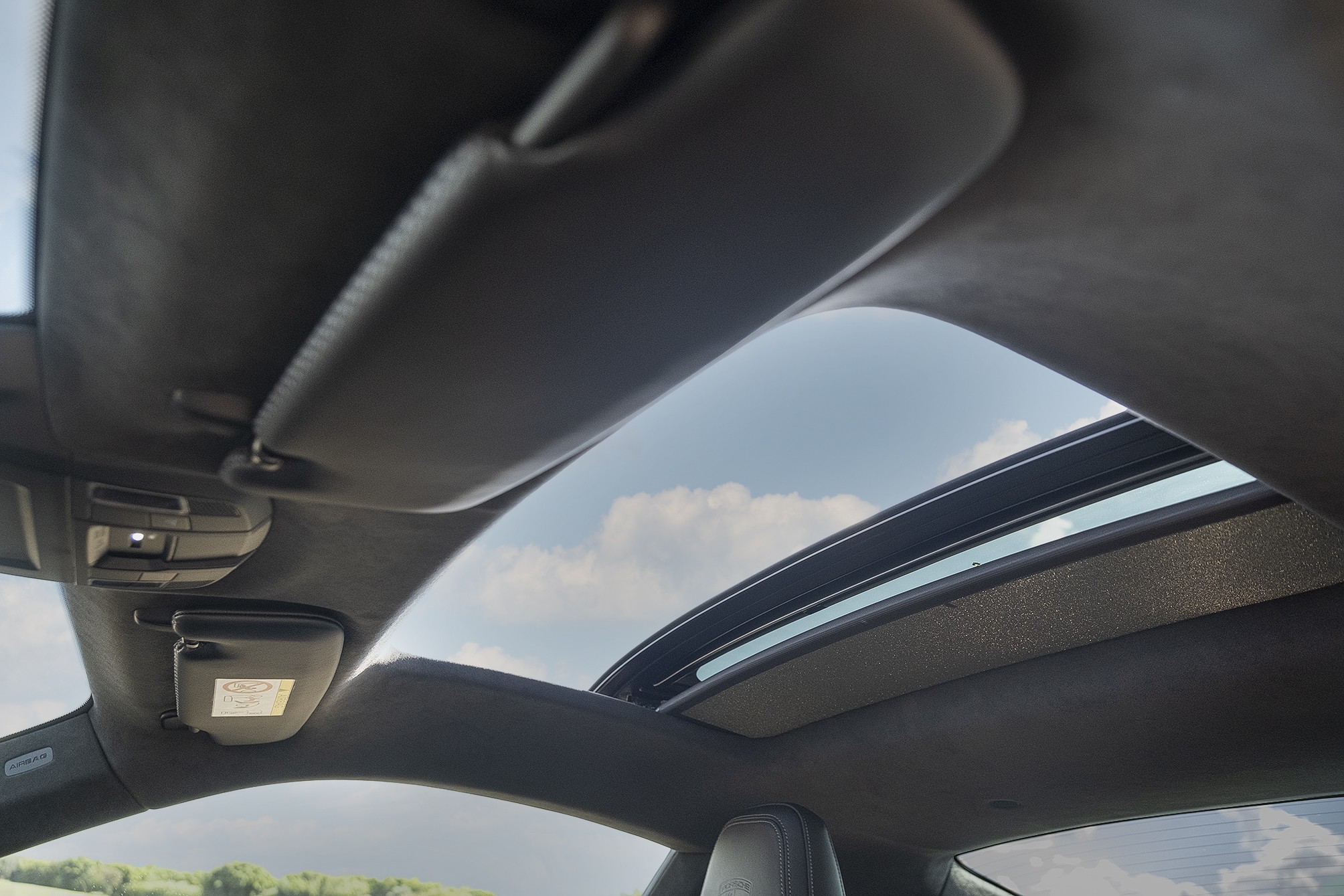 Interior de um carro com teto solar panorâmico aberto, mostrando o céu azul com nuvens brancas. O forro do teto é escuro, e os para-sóis estão levantados. À esquerda, um console de luzes internas está visível.