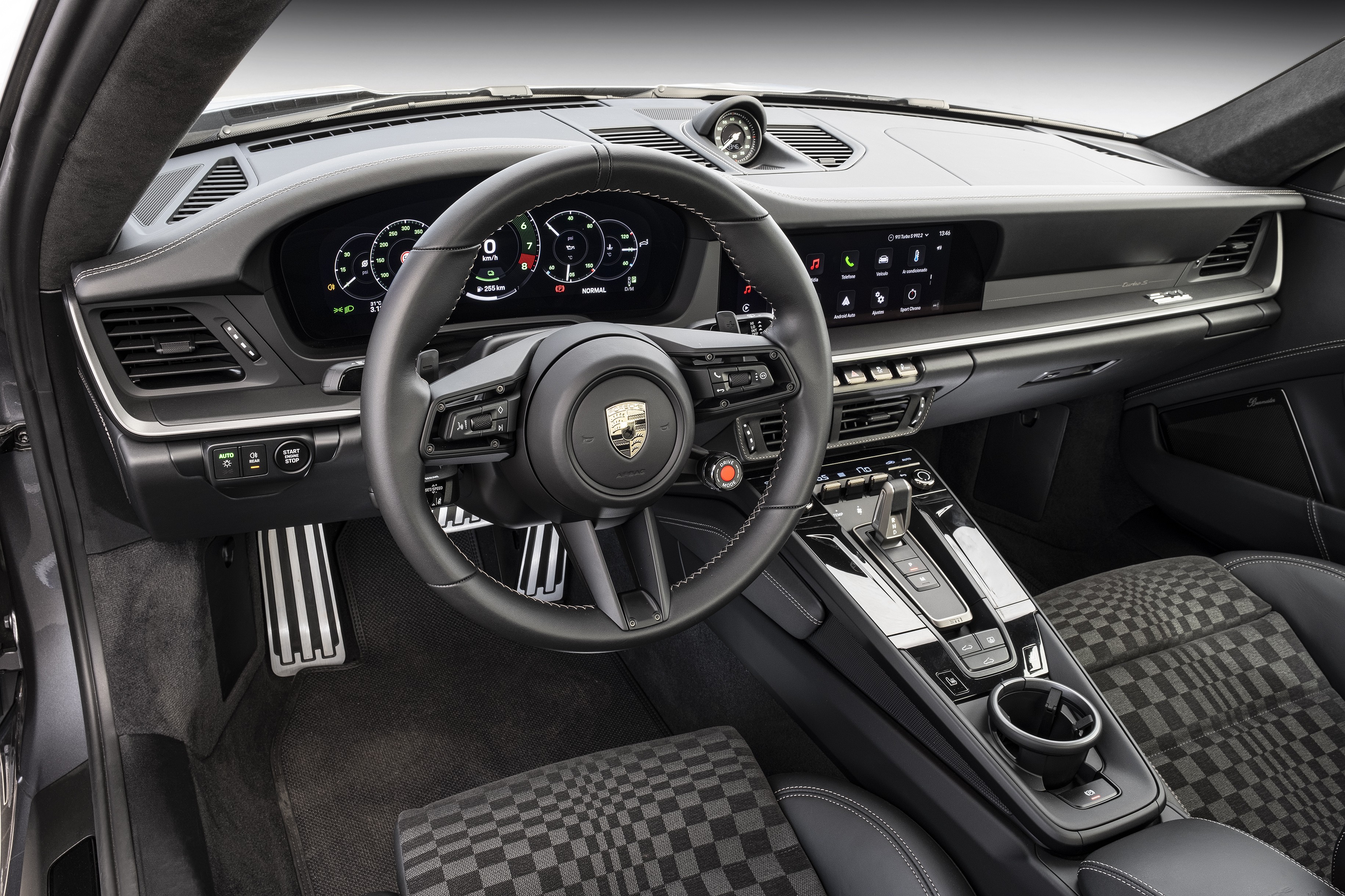 Interior de um carro esportivo de luxo, com volante multifuncional preto e o emblema da Porsche no centro. O painel digital exibe velocímetro e conta-giros. À direita, uma tela sensível ao toque com ícones de aplicativos. O console central tem alavanca de câmbio, botões de controle e porta-copos. Os bancos são pretos com estampa xadrez em tons de cinza. Pedais de metal escovado no chão. O ambiente é moderno e sofisticado