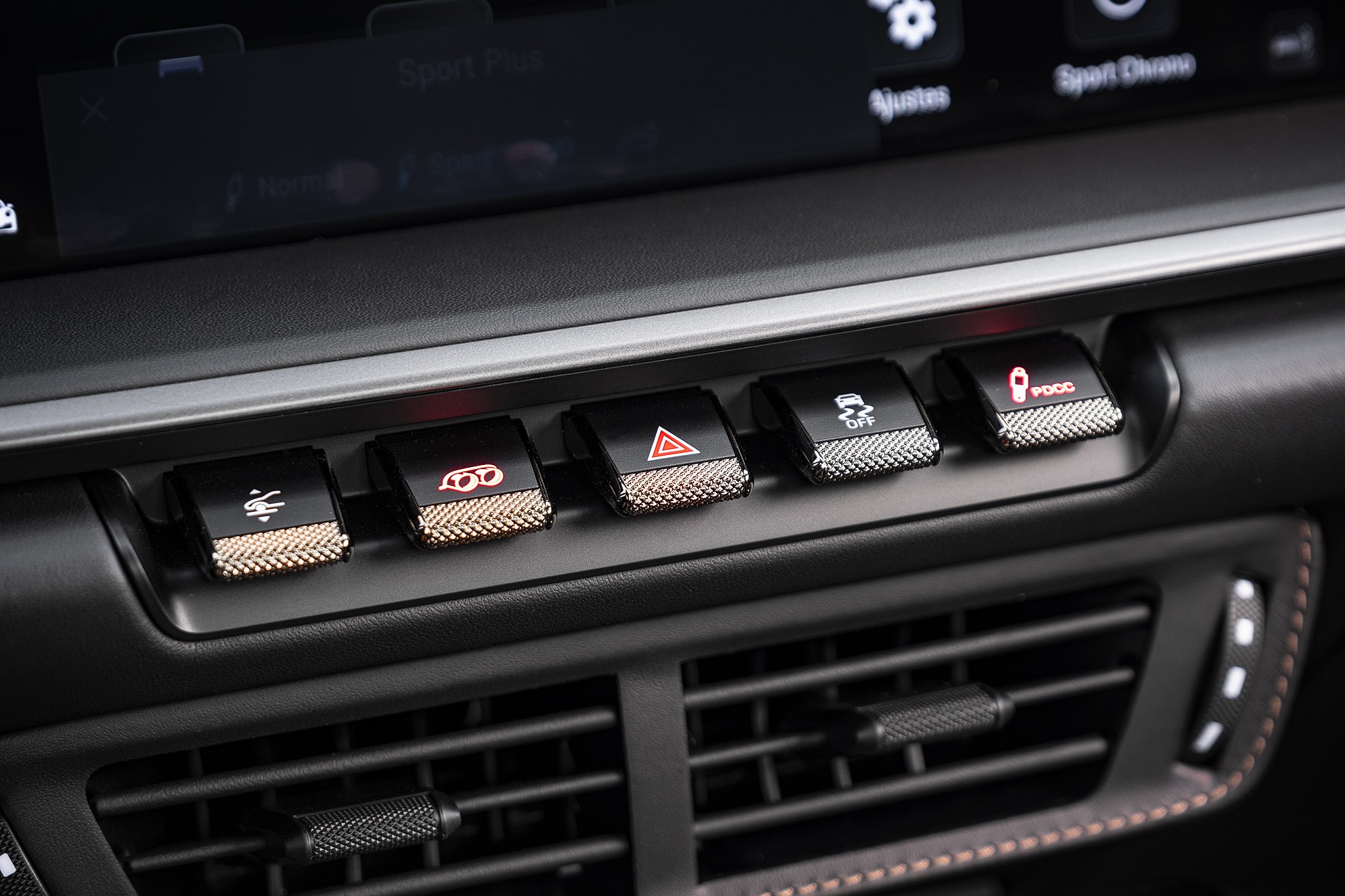 Painel de controle de carro com cinco botões pretos e texturizados em dourado ou prata, cada um com um ícone iluminado em vermelho ou branco, como um triângulo de alerta e um carro derrapando. Abaixo, saídas de ar pretas com detalhes em couro costurado.
