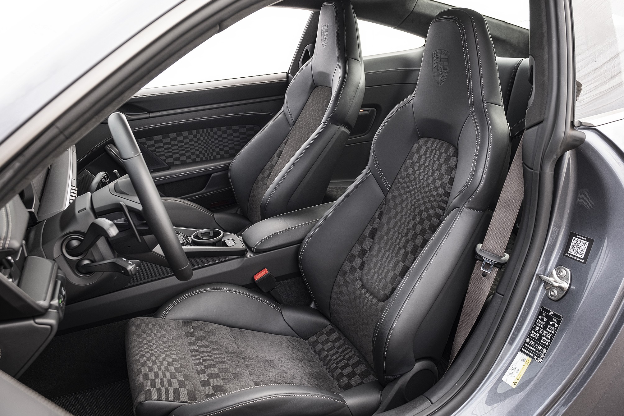 Interior de carro esportivo cinza com bancos de couro preto e tecido xadrez, volante e painel escuros, e cinto de segurança cinza claro