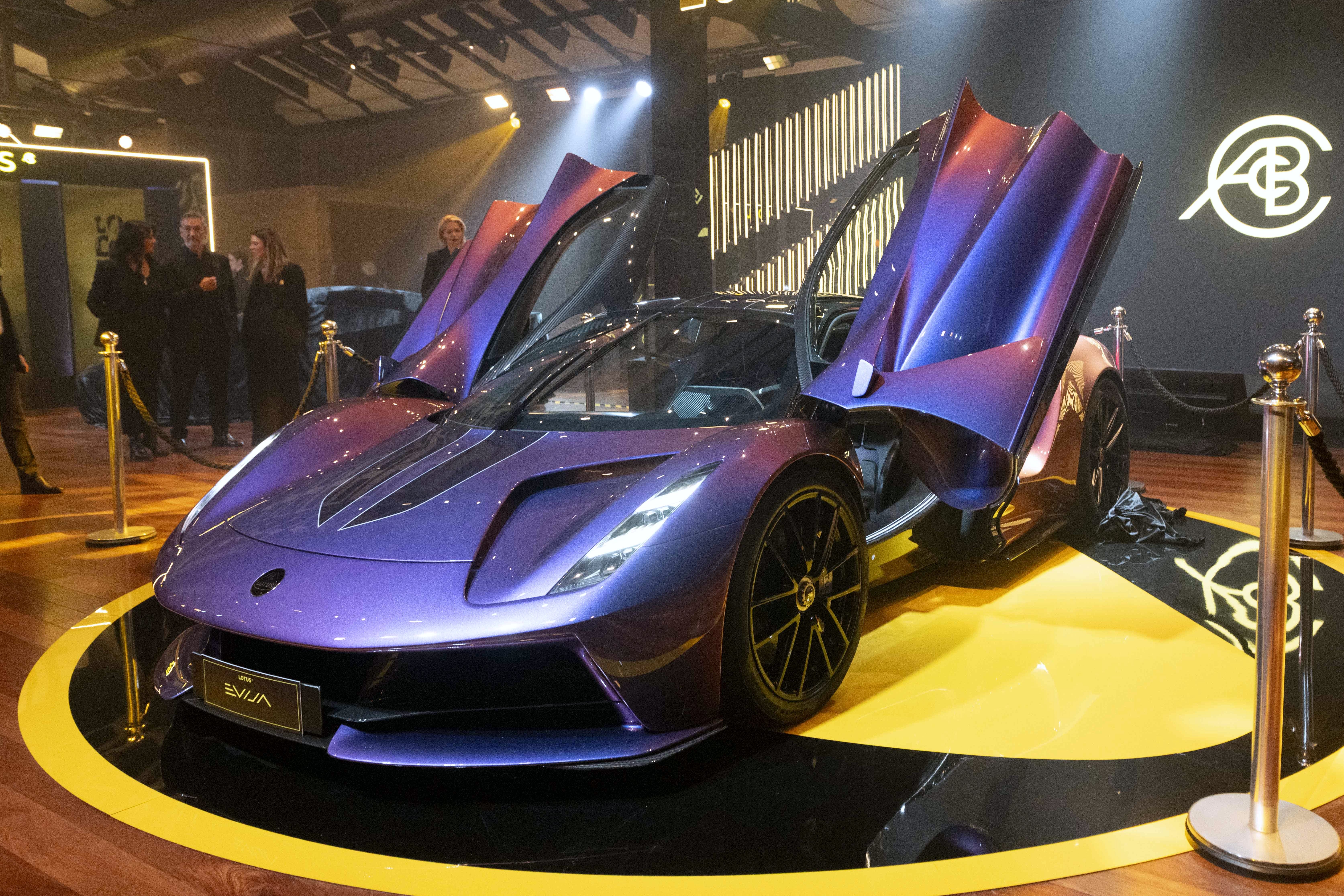 Carro esportivo roxo metálico com portas abertas para cima, exibido em plataforma preta e amarela. Pessoas observam ao fundo, em ambiente com iluminação focada e um logotipo branco na parede