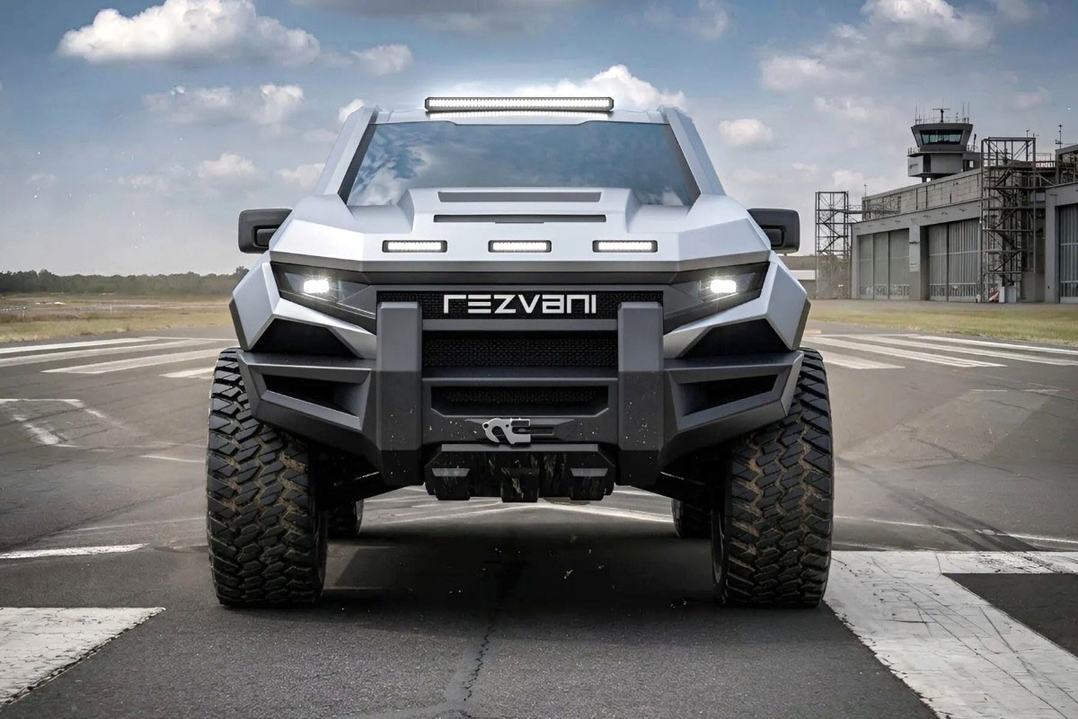 Caminhonete Rezvani Tank cinza-claro, com design robusto e futurista, para-choque dianteiro preto, faróis e barras de LED acesos, pneus off-road grandes, estacionada em uma pista de aeroporto com nuvens no céu e um hangar ao fundo