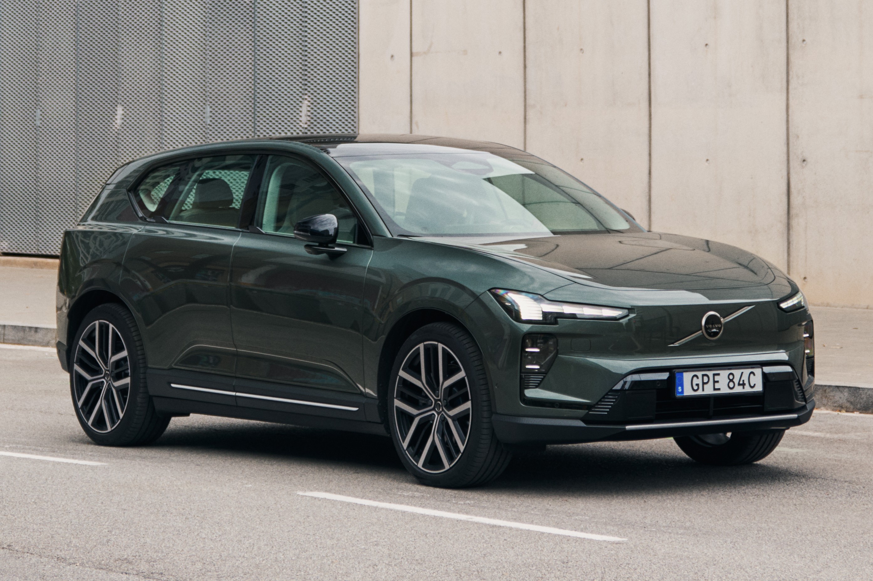 SUV elétrico Volvo EX30 verde escuro, visto de frente e lateral, parado em uma rua asfaltada com faixas brancas, em frente a uma parede cinza texturizada