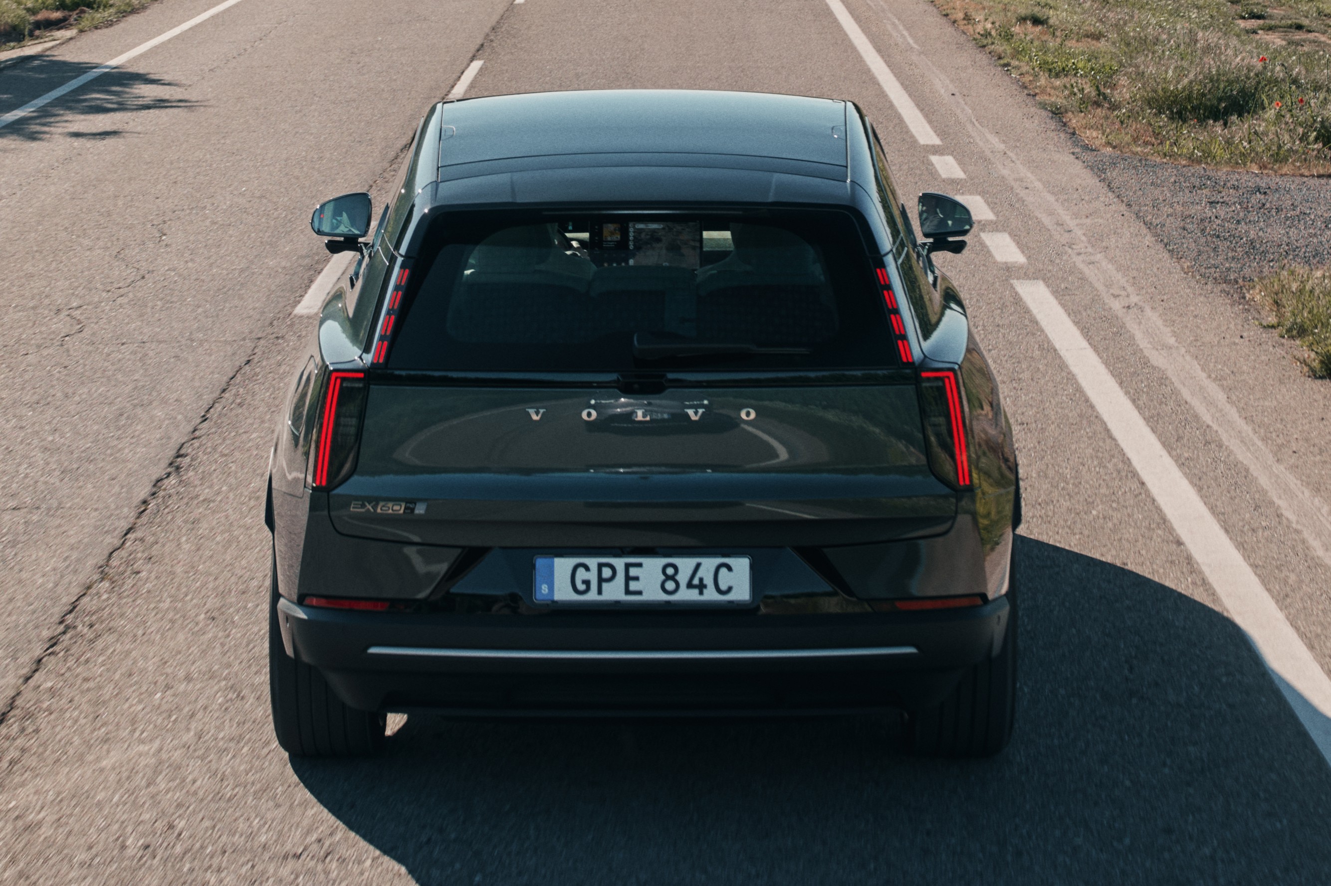 Traseira de um carro Volvo preto em uma estrada asfaltada com faixas brancas, sob luz solar, com vegetação seca à direita
