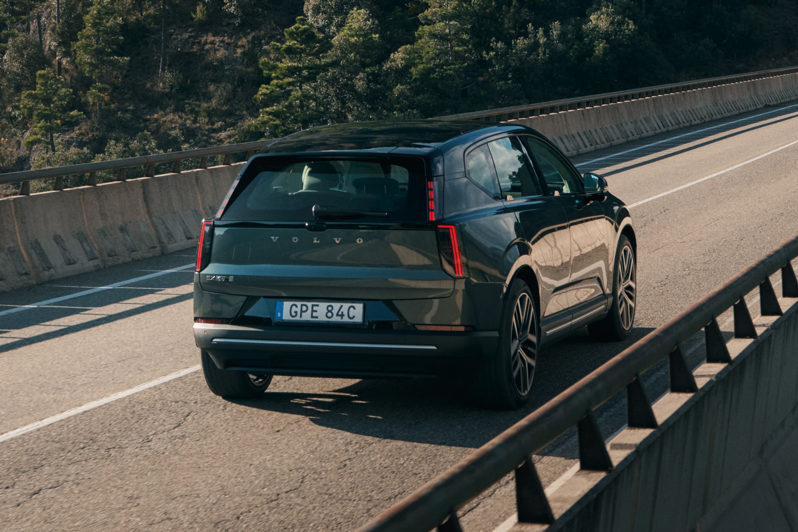 SUV Volvo EX90 escuro em uma estrada de asfalto, visto por trás, com montanhas arborizadas ao fundo