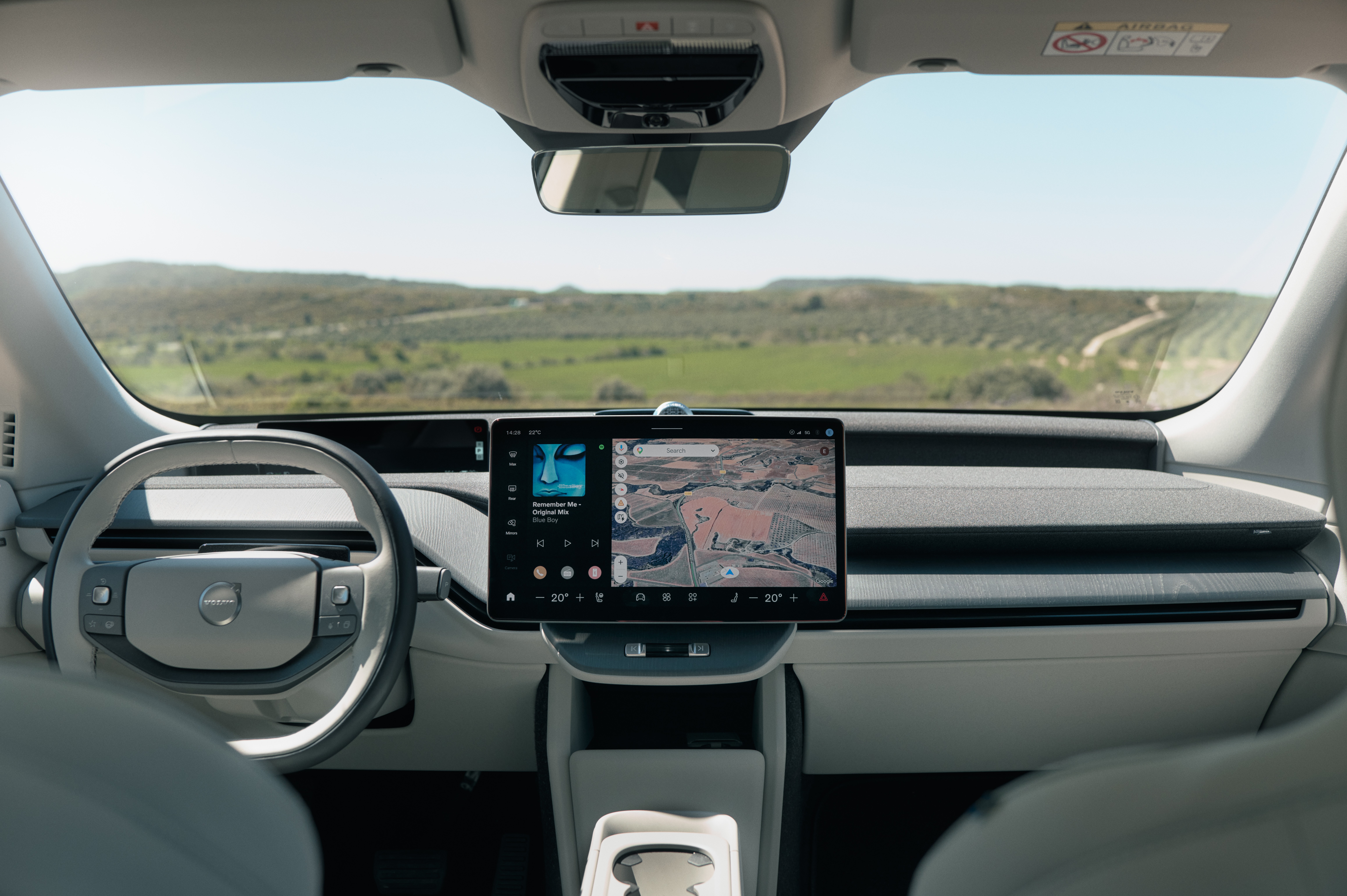 Interior de um carro moderno com volante cinza claro à esquerda e uma tela central exibindo um mapa e reprodutor de música. O para-brisa revela uma paisagem verde de colinas sob céu azul