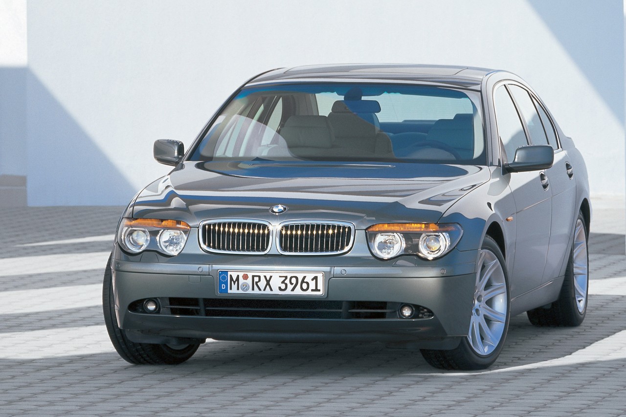 Carro BMW Série 7 cinza escuro estacionado em uma superfície de blocos, com uma parede branca ao fundo e sombra projetada