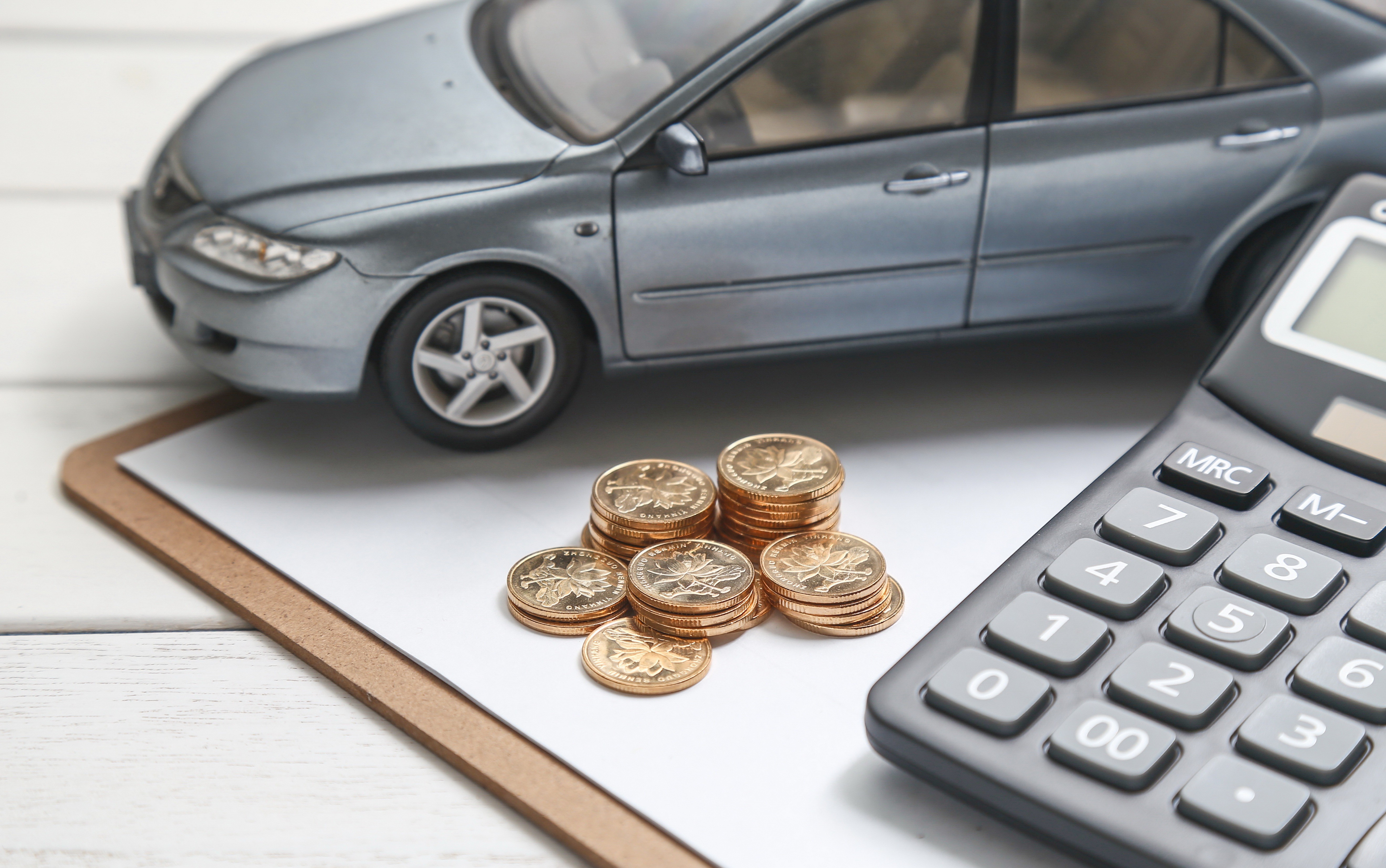 Miniatura de carro cinza sobre prancheta branca, com pilhas de moedas douradas e uma calculadora preta à direita, simbolizando custos veiculares