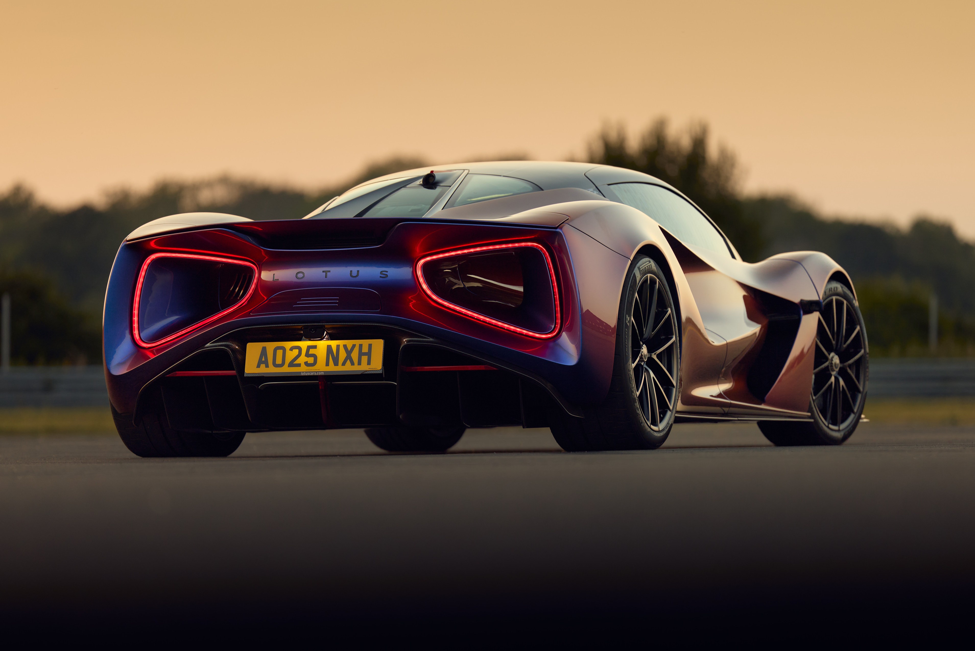 Traseira de um carro esportivo Lotus Evija roxo e vermelho, com luzes traseiras em LED vermelhas acesas, parado em uma pista de asfalto sob um céu alaranjado ao pôr do sol