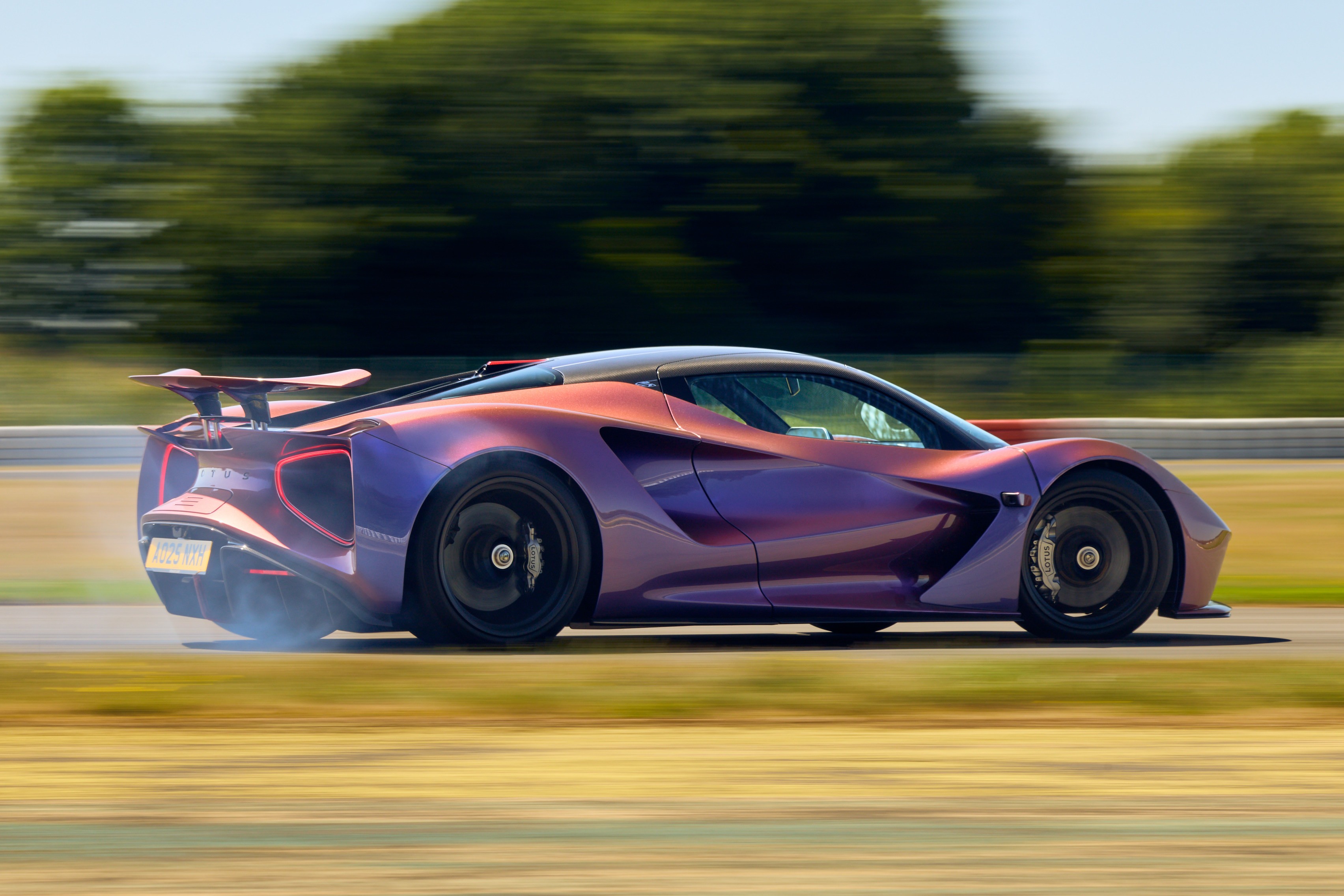 Carro esportivo roxo e cobre em alta velocidade, com fumaça saindo do pneu traseiro, em uma pista de corrida com árvores borradas ao fundo