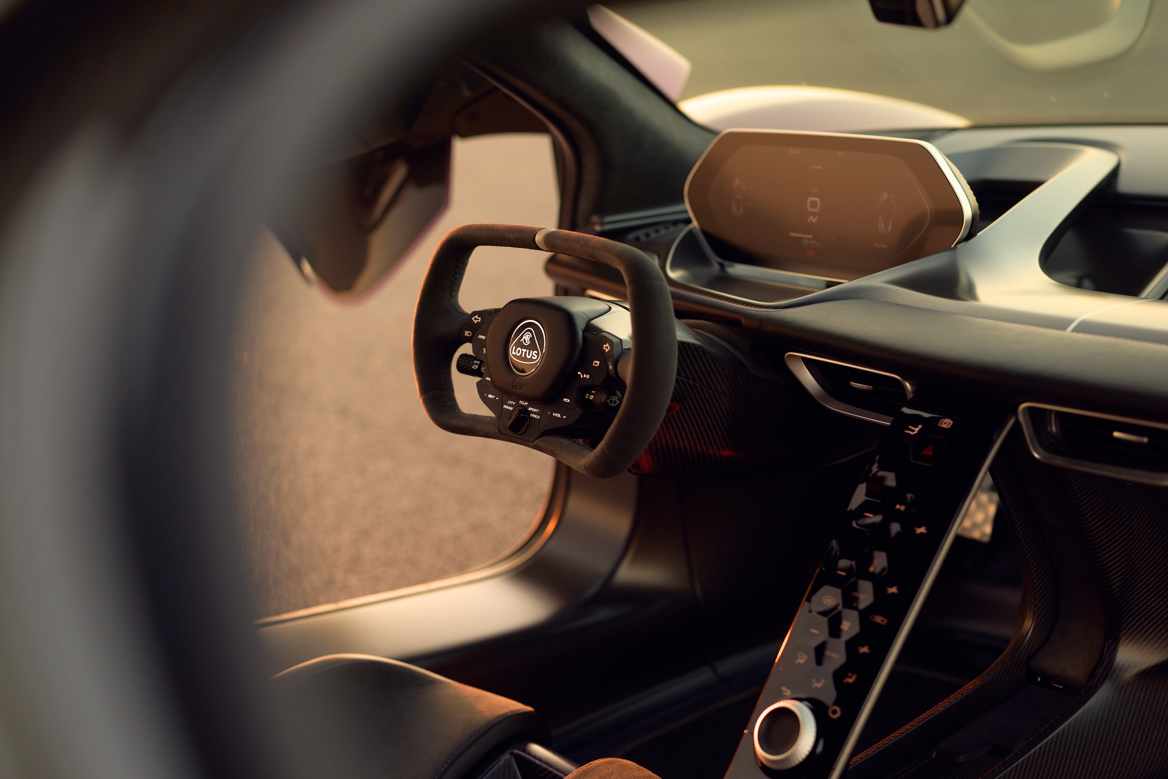 Interior de um carro esportivo Lotus, com volante preto de três raios e o logotipo da marca ao centro. À direita, um painel digital retangular e um console central com botões hexagonais e um seletor circular. O ambiente é escuro, com detalhes em cinza e iluminação suave