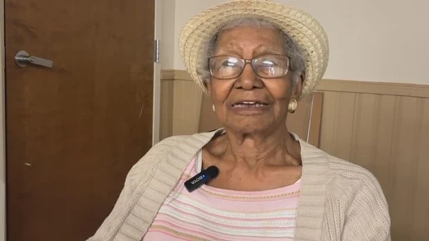 Mulher idosa, pele escura, cabelos grisalhos, óculos e chapéu de palha, usando blusa listrada rosa e cardigan bege, com microfone de lapela preso à blusa, sorrindo levemente, em ambiente interno com porta marrom ao fundo