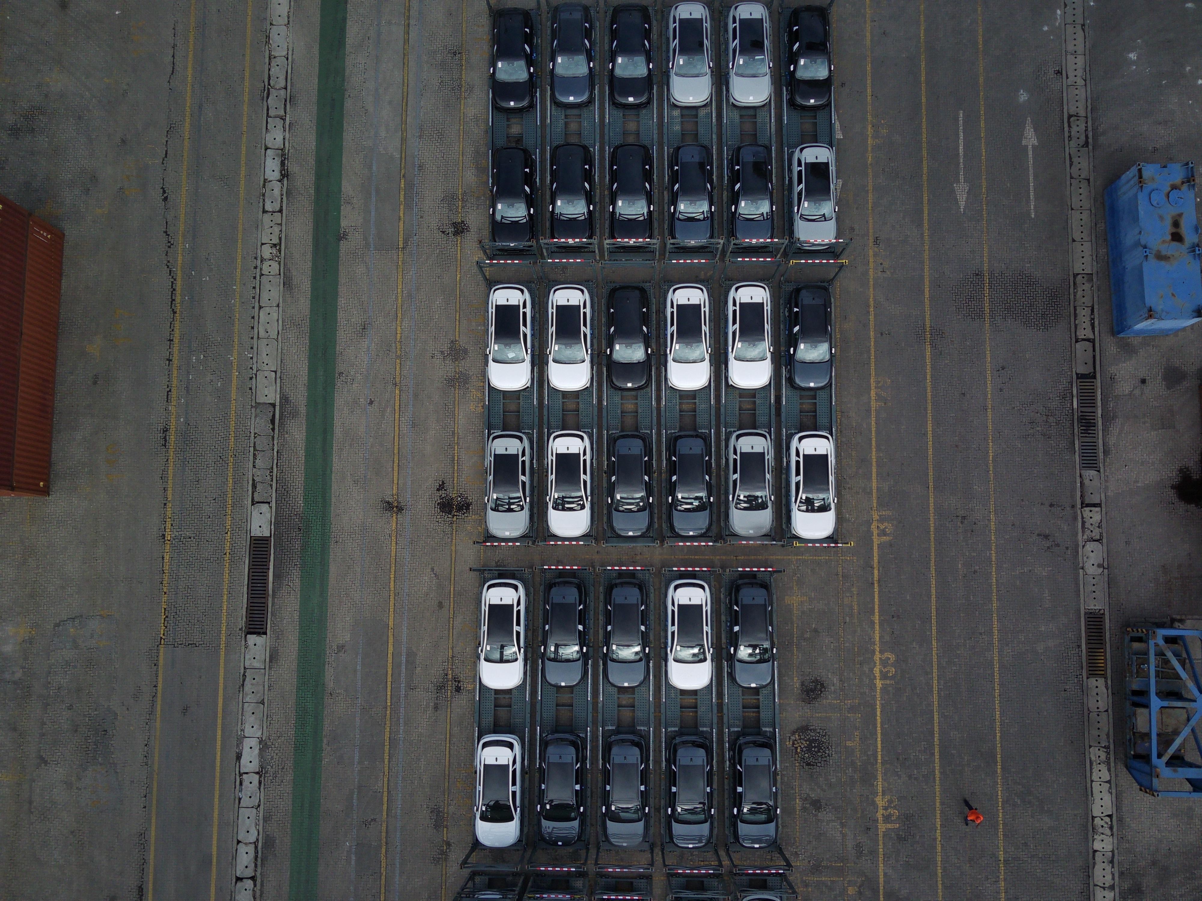 Vista aérea de dezenas de carros novos, brancos e pretos, estacionados em fileiras sobre plataformas de transporte em um pátio de concreto. À esquerda, um contêiner marrom e à direita, um contêiner azul. Um trabalhador de laranja caminha no canto inferior direito.
