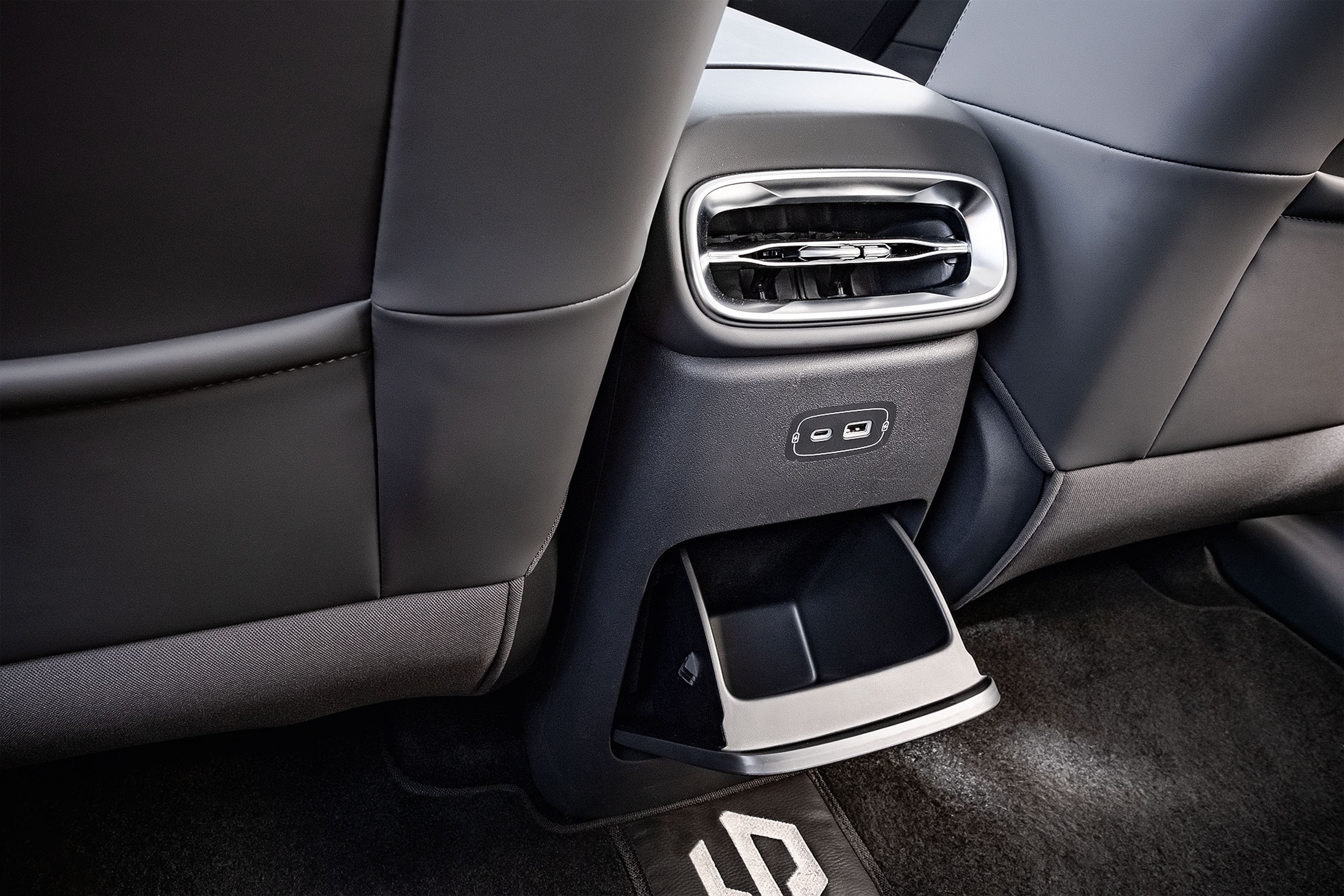Interior de carro com bancos traseiros de couro preto, console central com saídas de ar cromadas, duas portas USB-C e um porta-objetos retrátil, sobre tapete escuro com logo